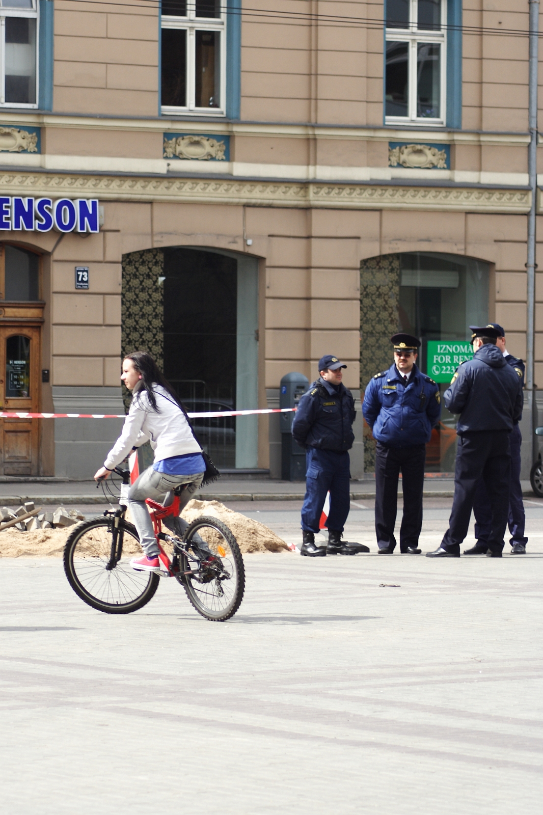 A woman rides a bicycle past a group of four police officers standing beside a red and white caution tape. They appear to be securing a construction area marked by the tape and some soil on the ground. The scene, reminiscent of Kritiska masa, unfolds in front of a beige building with green and blue trim around the windows.