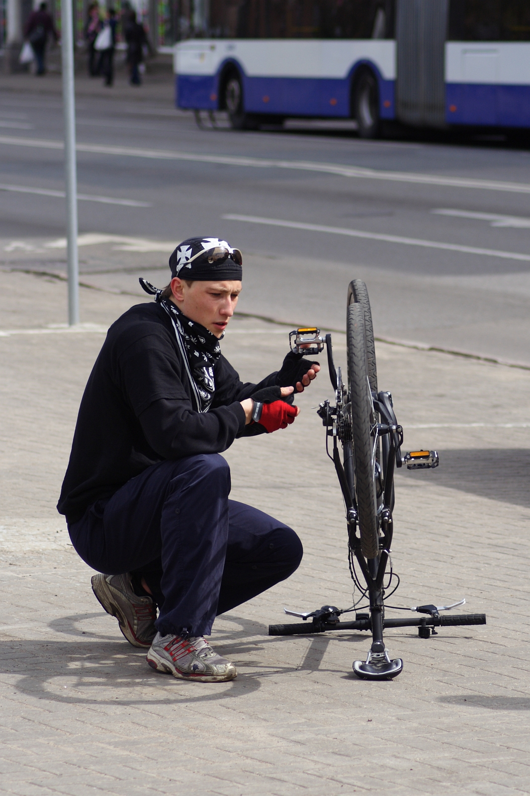 A person wearing a bandana, gloves, and a black sweatshirt is kneeling and working on an upside-down bicycle on a sidewalk during the Kritiska masa event in 2010. The background shows an unfocused street with a white and blue bus. The individual appears focused on adjusting the bike's pedal or chain.
