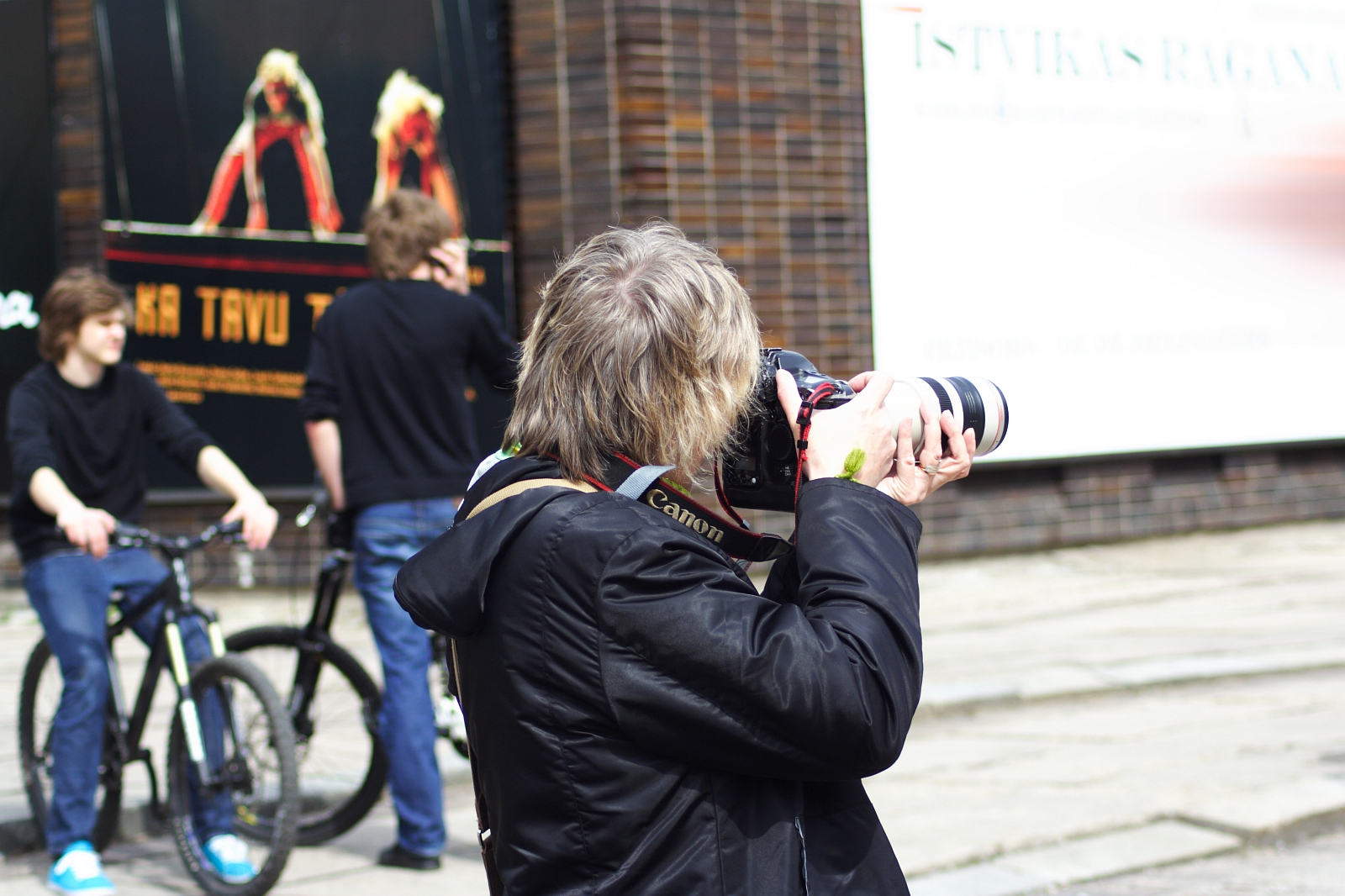 A person with gray hair, wearing a dark coat, photographs something with a large Canon camera attached to a telephoto lens. In the background of this 2010 scene, two young people with bikes are visible—one standing and one sitting—alongside a large poster and part of a building facade, perhaps hinting at Kritiska masa.