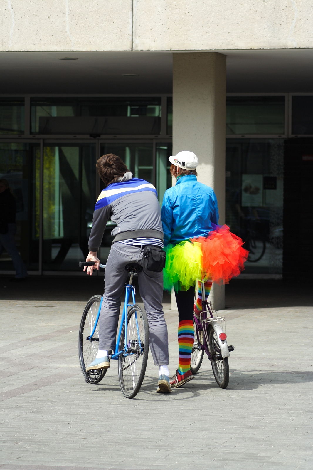 Two bicyclists in front of a building during the 2010 Kritiska masa event. The person on the left is wearing a gray outfit with blue stripes, while the person on the right is dressed in colorful clothing, including a blue jacket, neon tutu, and rainbow-striped leg warmers. Both are looking forward, their backs facing the camera.