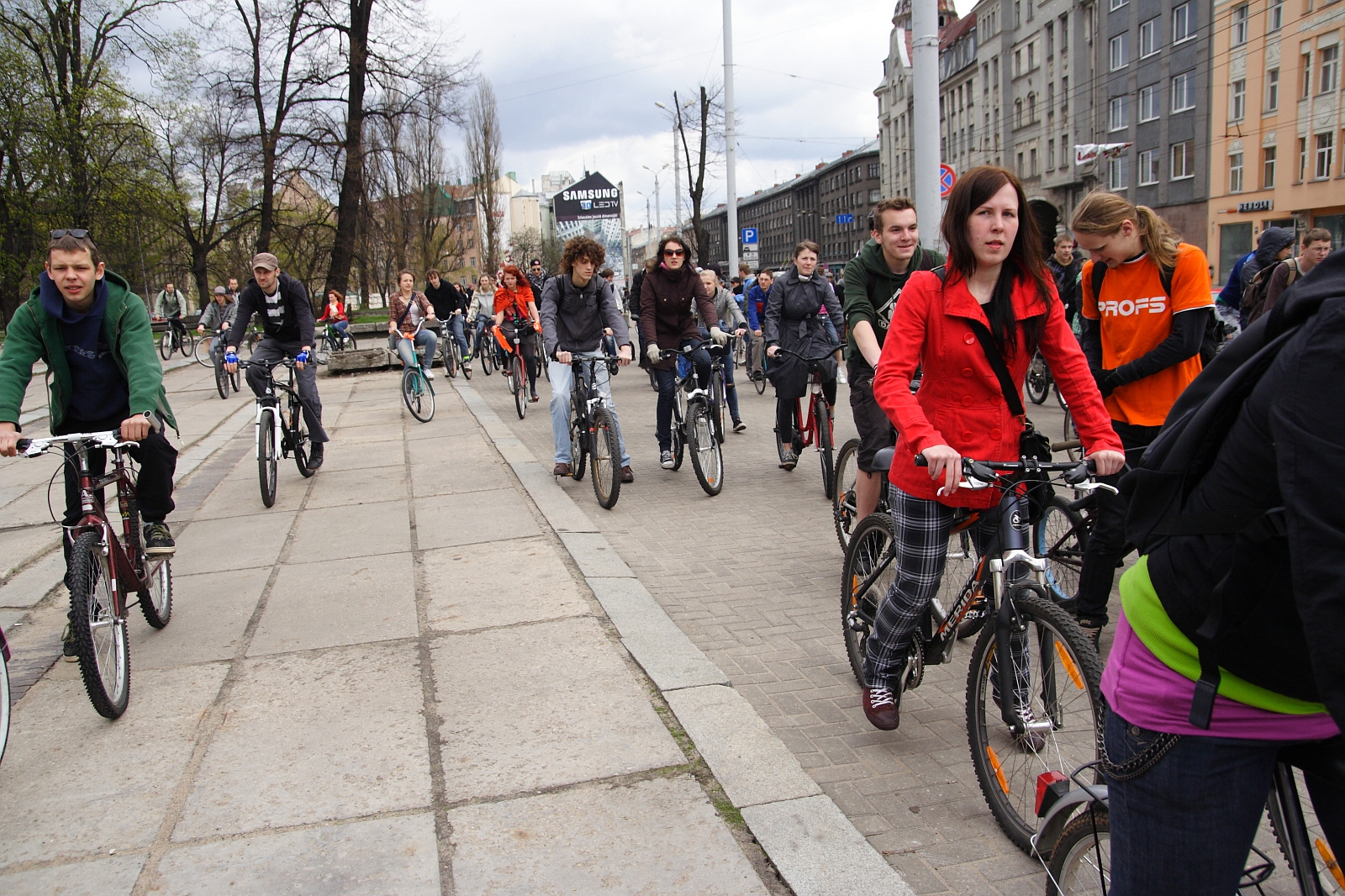 A diverse group of people, some dressed casually and others in sporty attire, ride bicycles on a city street during the 2010 Kritiska masa event. Lined with buildings and marked by trees in the background, the cloudy weather only enhances the scene's active and communal atmosphere.