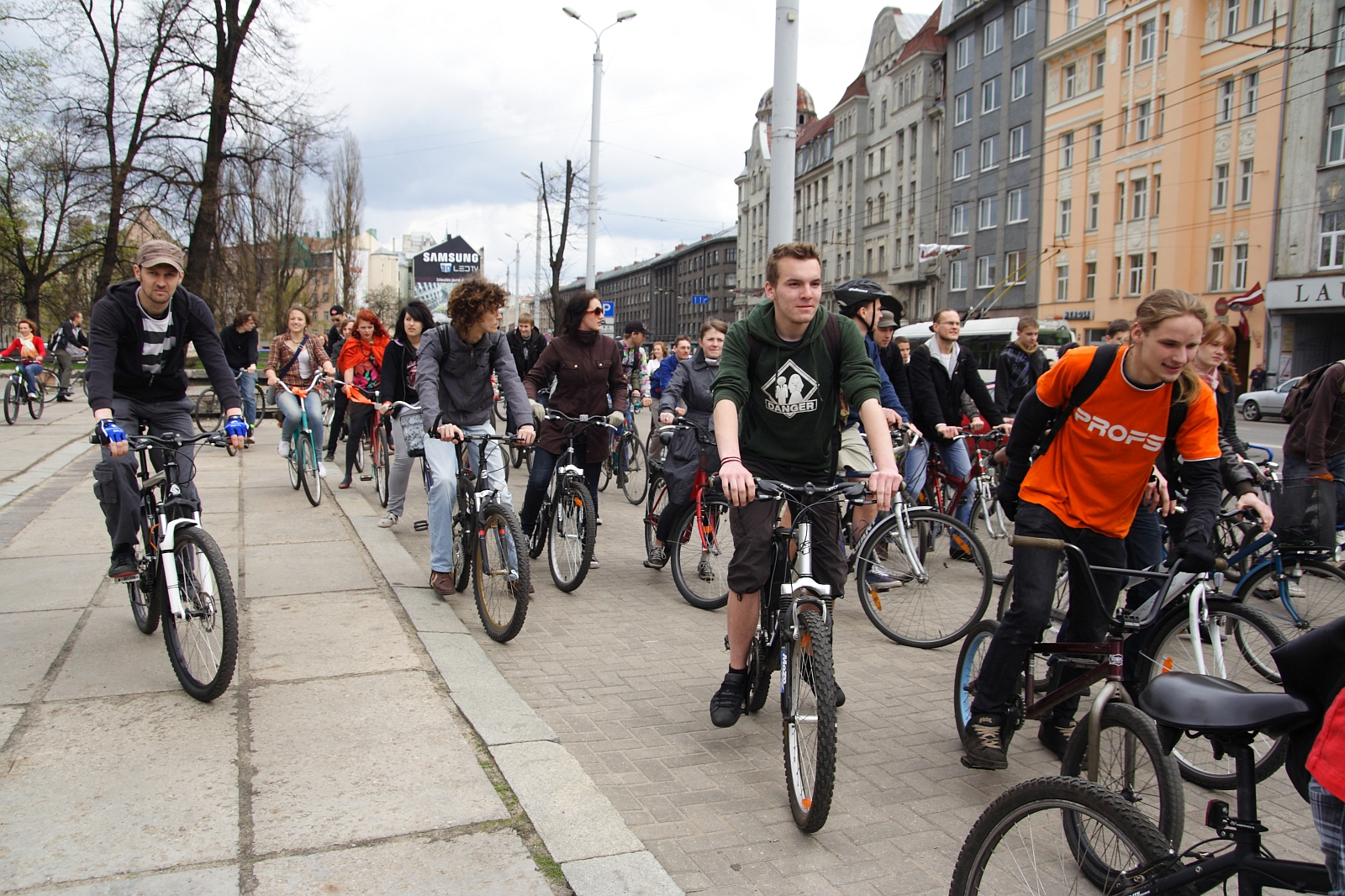 A large group of people of various ages is participating in a Kritiska masa bike ride along a city street. They are riding bicycles and appear to be in good spirits, wearing casual clothing. The backdrop features tall, colorful buildings and a partially cloudy sky, capturing the essence of this 2010 community event.