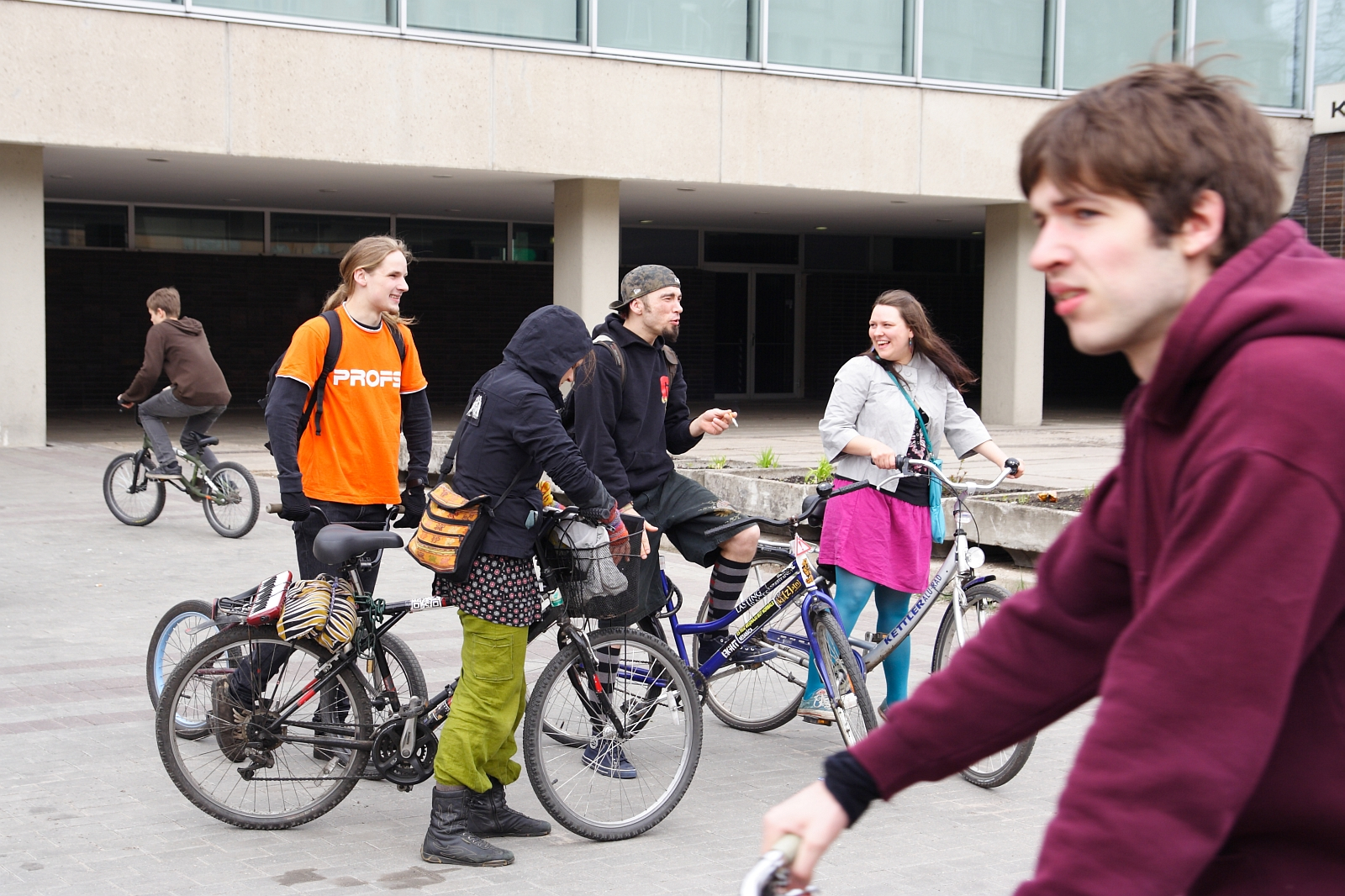 A group of five young adults stands together with their bicycles in an urban area, engaging in conversation. One person in focus rides a bike in the foreground. The background features a large building with glass windows, recalling the 2010 Kritiska masa vibes. The weather appears overcast, and everyone is dressed casually.