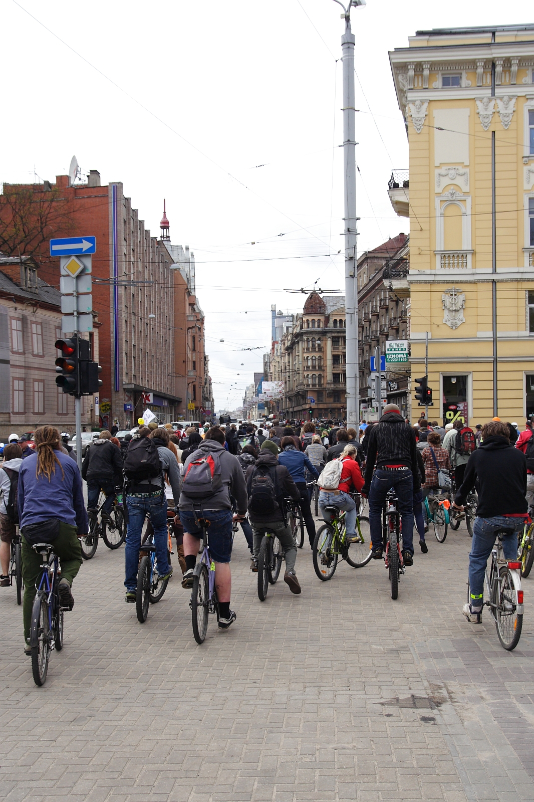 A large group of cyclists rides down a city street bordered by both modern and historic buildings. The street, equipped with traffic lights and street signs, shows an overcast sky above. The cyclists, some wearing helmets, travel closely together in what looks like a Kritiska masa 2010 community ride or event.