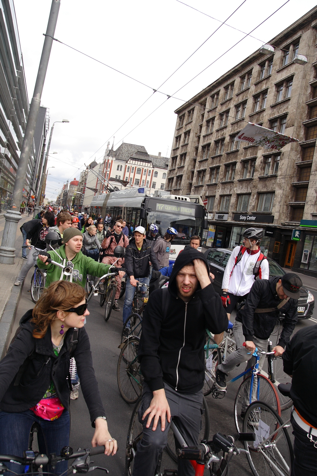 A large group of cyclists in casual attire ride along a city street, surrounded by tall, old buildings. Some cyclists wear helmets while others do not. The atmosphere appears busy and urban, reminiscent of the 2010 Kritiska Masa event, with a mix of buses and other structures in the background. The cyclists seem focused on their travel.