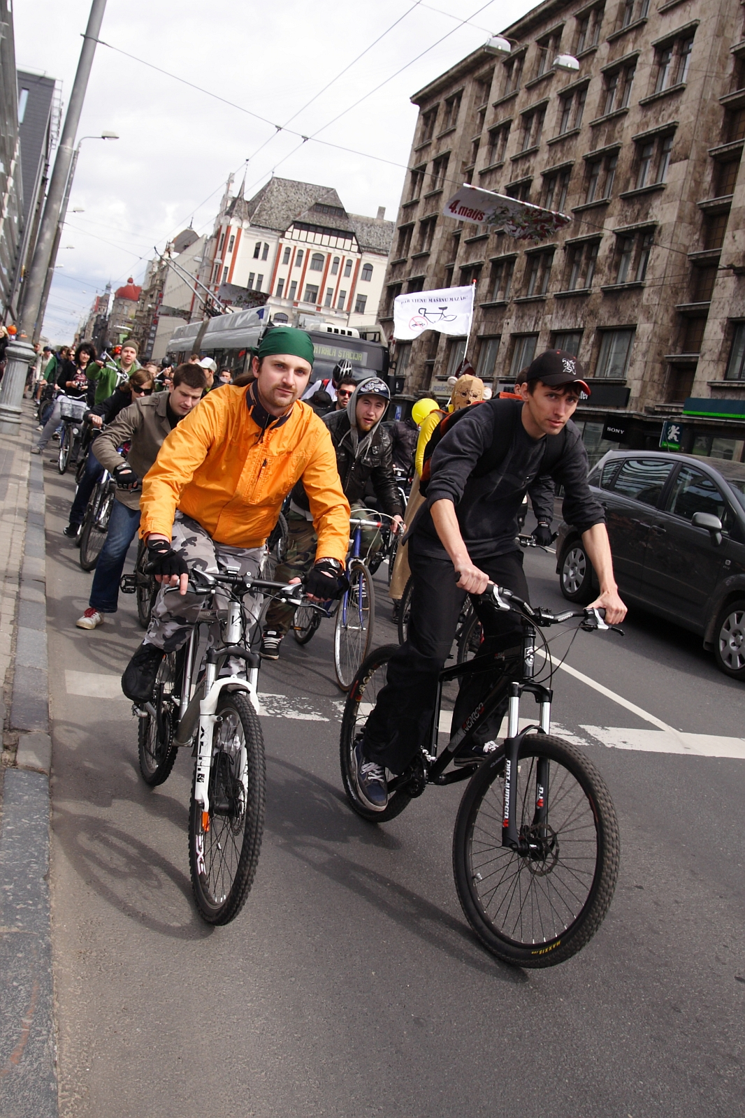 A group of cyclists is riding on a city street, reminiscent of the Kritiska masa event in 2010. Two men in the forefront are wearing an orange jacket and a black jacket, respectively. Behind them, more cyclists follow, with buildings lining both sides of the street. Some pedestrians can also be seen on the sidewalk to the left.