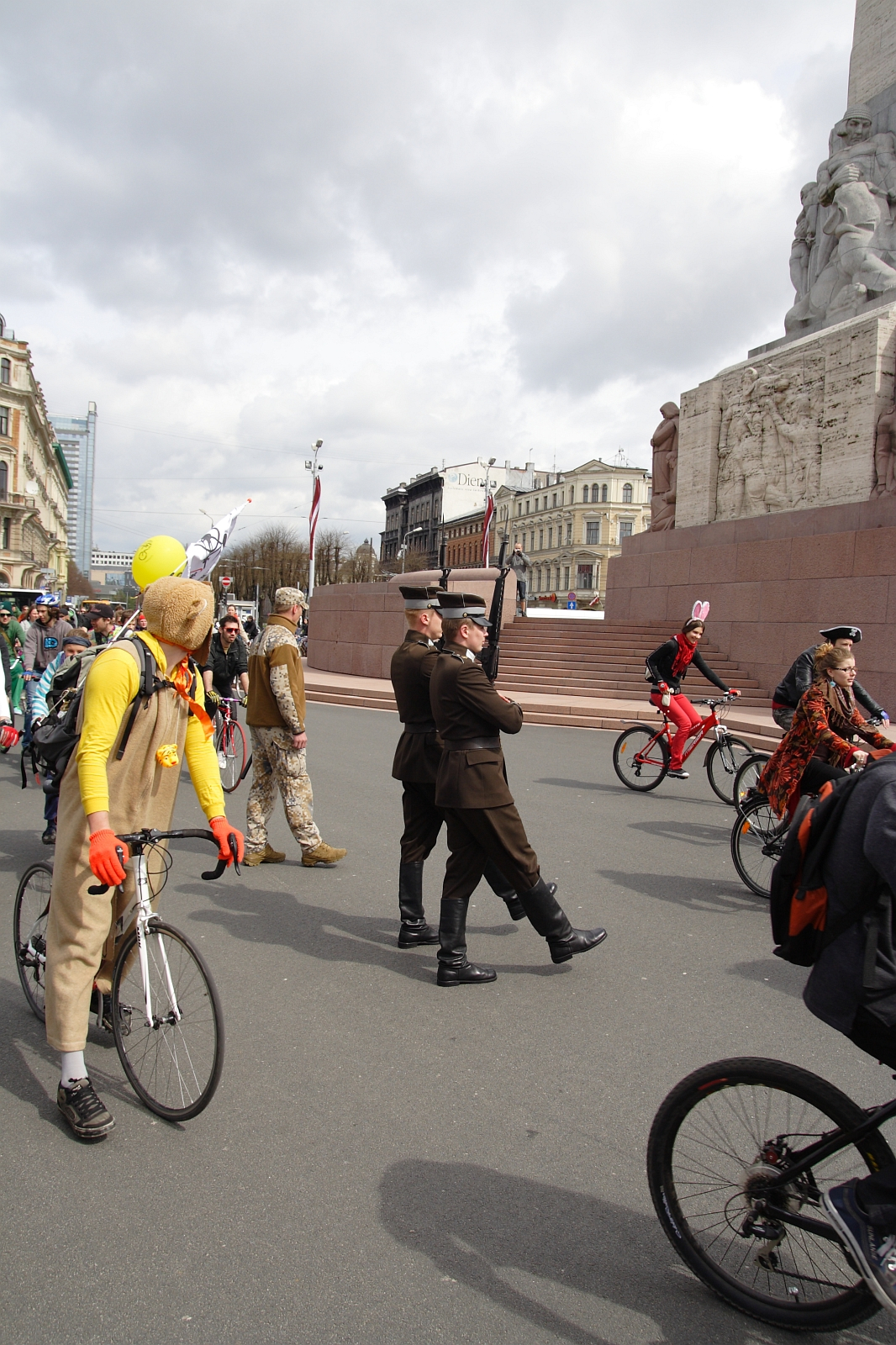 A group of people participates in a quirky cycling event, Kritiska masa 2010. Cyclists in colorful and eccentric costumes ride near a large monument. Two men in military uniforms stand in the center, one saluting. Balloons and flags add to the festive atmosphere against a backdrop of historical buildings.
