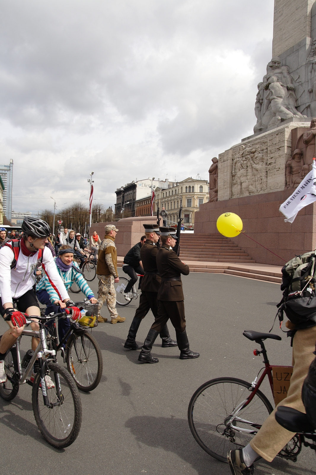 A busy urban scene in 2010 features a mix of cyclists, pedestrians, and onlookers in front of a large monument. People are casually dressed, some are pushing strollers, and one person is holding a yellow balloon. The sky is overcast, and buildings can be seen in the background, reminiscent of Kritiska masa events.