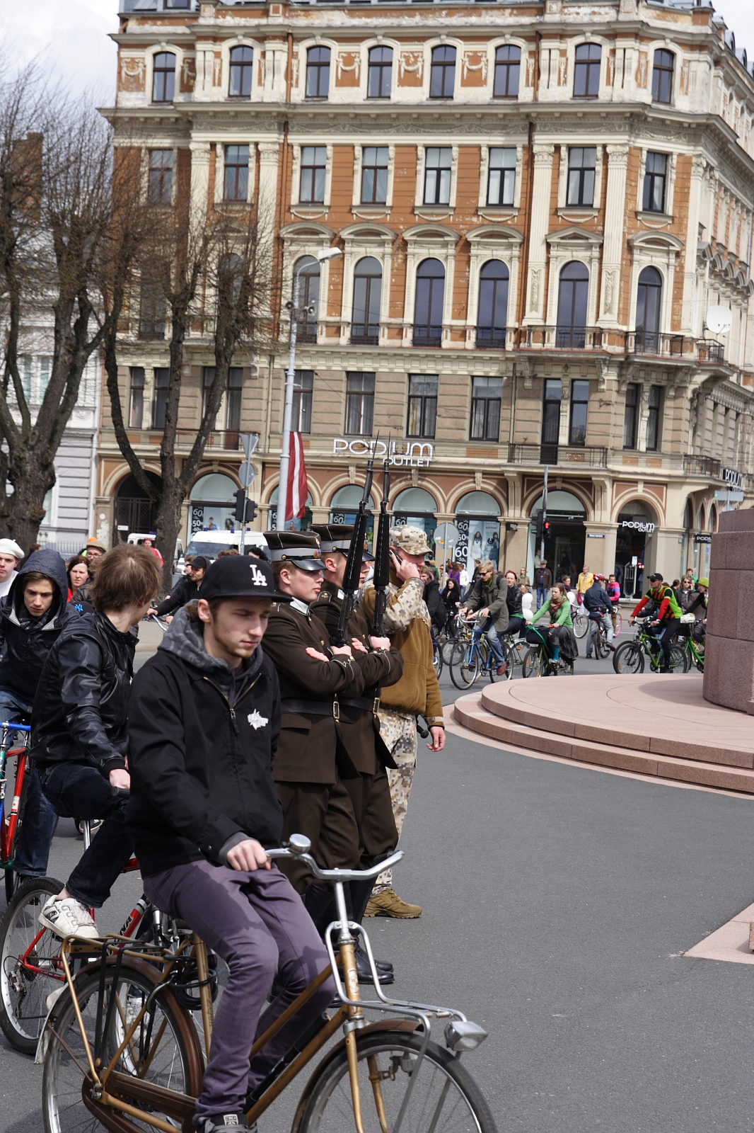 A group of cyclists from the Kritiska masa rides past a military ceremony in front of an ornate, historic building. Soldiers in various uniforms, including camouflage, are standing at attention while holding rifles. Spectators and additional cyclists are visible in the background. The sky is overcast, reminiscent of events from 2010.