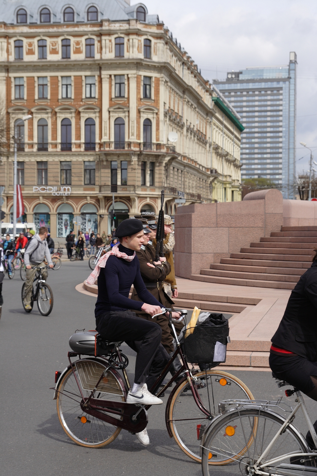 People ride bicycles on a city street near a grand historic building with ornate architecture and large windows. In the foreground, a person wearing a black coat and a beret cycles past, reminiscent of the 2010 Kritiska masa events, while others ride bikes in the background. A modern glass skyscraper is visible in the distance.