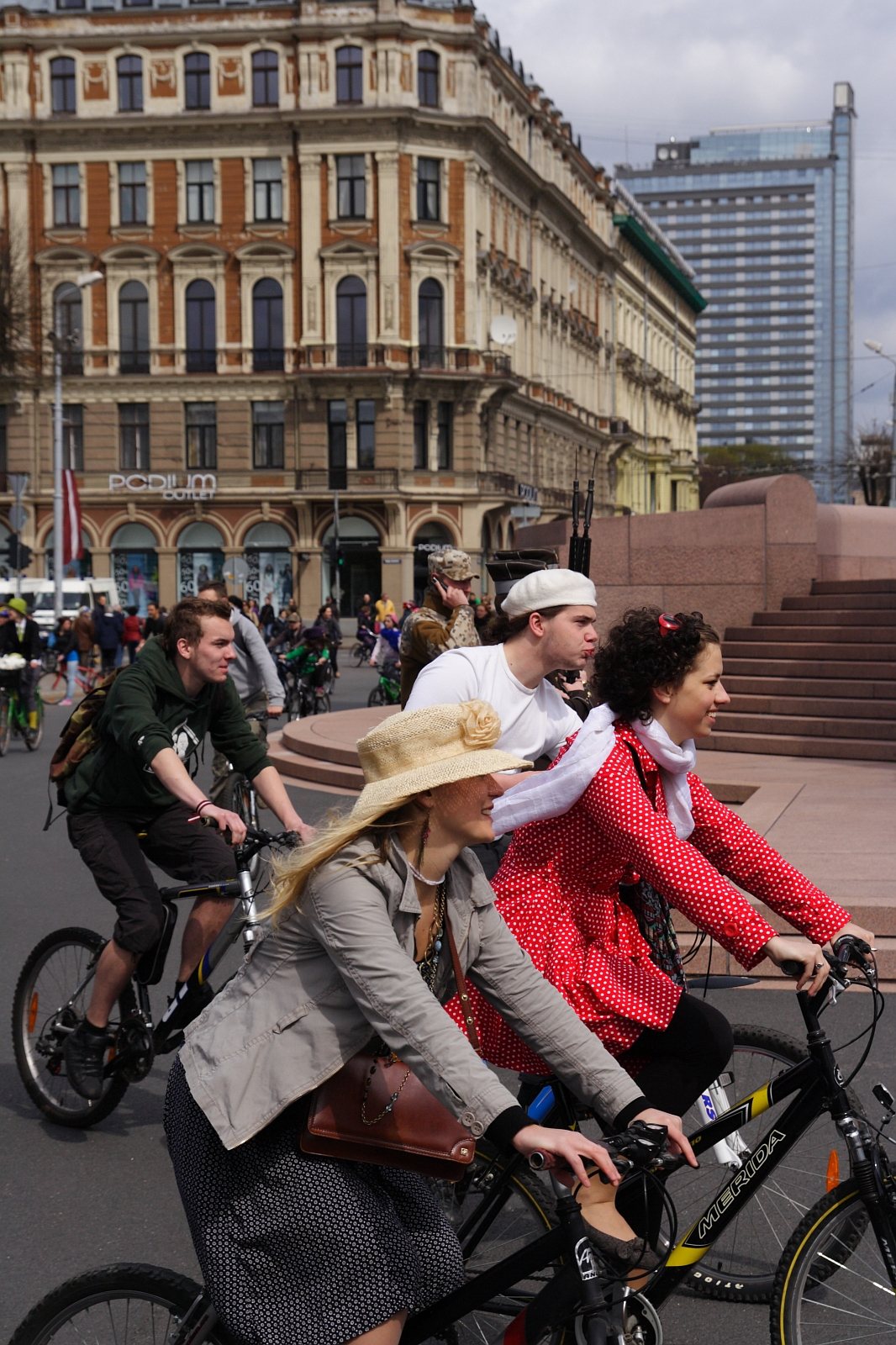 A group of people enjoy a bike ride through a city street during the 2010 Kritiska masa event. The joyful riders feature a man in a green hoodie, another in a pirate costume, a woman in a red polka-dot dress, and a woman in a beige hat and jacket. In the background are historic and modern buildings.