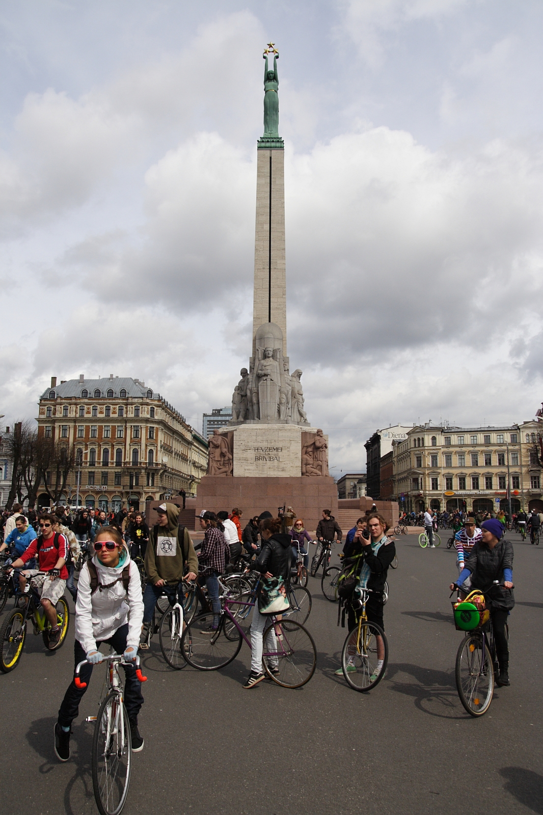 A large group of cyclists gathers in front of the Freedom Monument in Riga, Latvia for Kritiska masa 2010. The monument is tall with a statue of a woman holding three stars at the top. The cyclists, wearing casual clothing and helmets, are chatting and preparing to ride. Historic buildings and a cloudy sky are in the background.