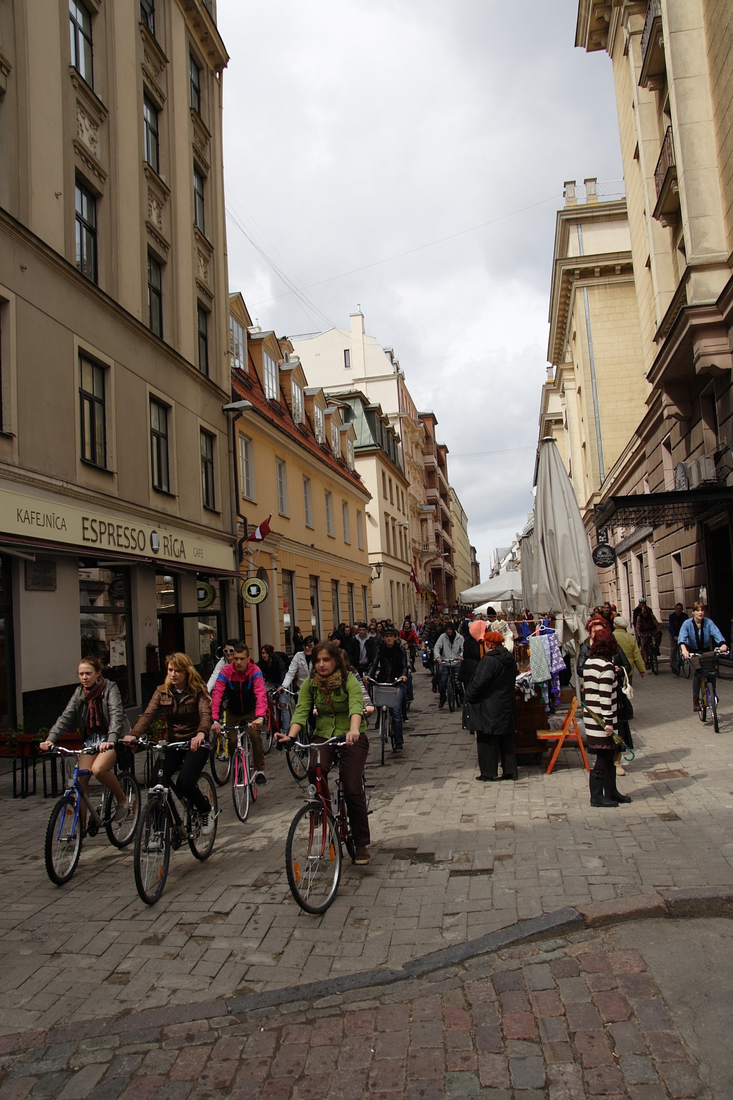 A bustling European street with historic buildings on both sides. Numerous pedestrians and cyclists navigate the cobblestone pavement, reminiscent of Kritiska masa 2010. A café named "Kafejnica ESPRESSO RIGA" is visible on the left. Various stalls and umbrellas line the right side, indicating a street market. The sky is overcast.