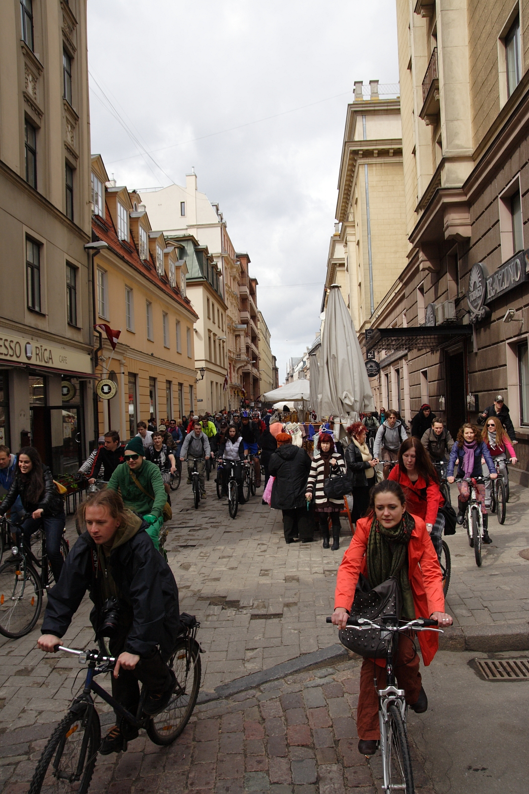 A busy, narrow street in a European city is filled with people riding bicycles and walking. The street, reminiscent of the 2010 Kritiska masa movement, is lined with tall historic buildings mixed with modern storefronts. Pedestrians and cyclists in vibrant clothing move actively under a cloudy sky, creating a lively atmosphere.