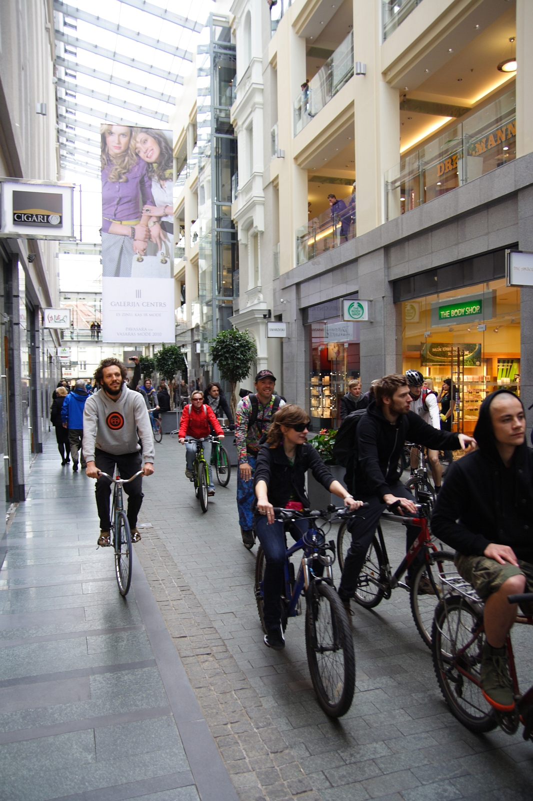 A group of people is riding bicycles through a modern, indoor shopping arcade with a glass ceiling, reminiscent of the 2010 Kritiska masa event. They are passing various stores, including a clothing store and a grocery store. Shoppers and pedestrians are visible in the background while a large advertisement banner hangs from the ceiling.