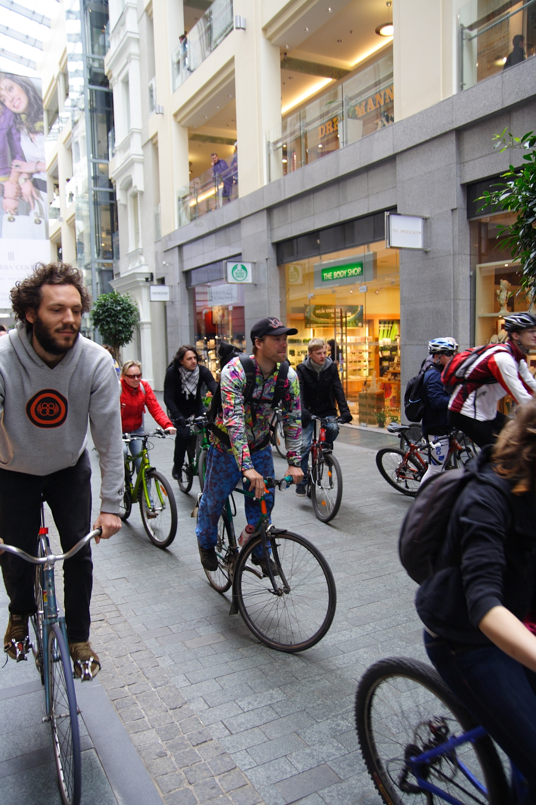 A group of cyclists from the 2010 Kritiska masa rides through a modern indoor shopping space. The cyclists wear varied attire: one in a gray hoodie with an orange emblem, and another in a colorful, patterned outfit. Shoppers and storefronts, including "The Body Shop," are visible in the background.