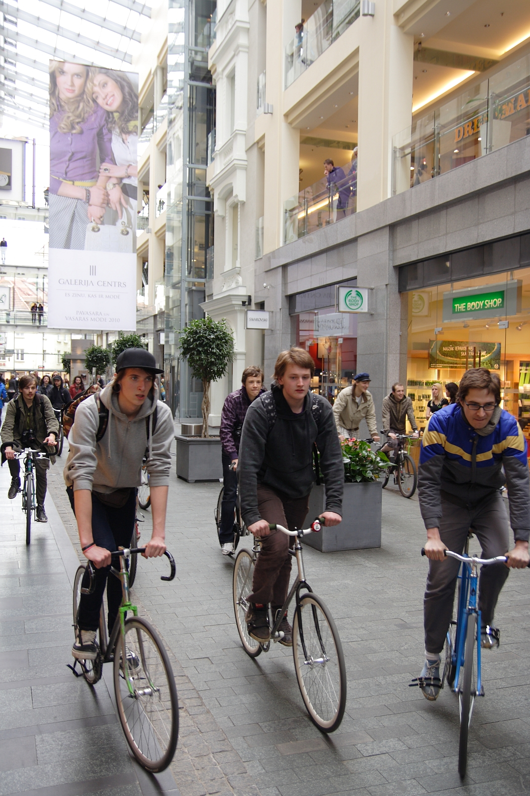Three young men are riding bicycles through an indoor shopping mall. The mall is bustling with pedestrians, and stores like The Body Shop are visible. In the background, a large vertical banner displays the words "Galleria Center," featuring an image of two women with stylish attire, reminiscent of a Kritiska masa event from 2010.