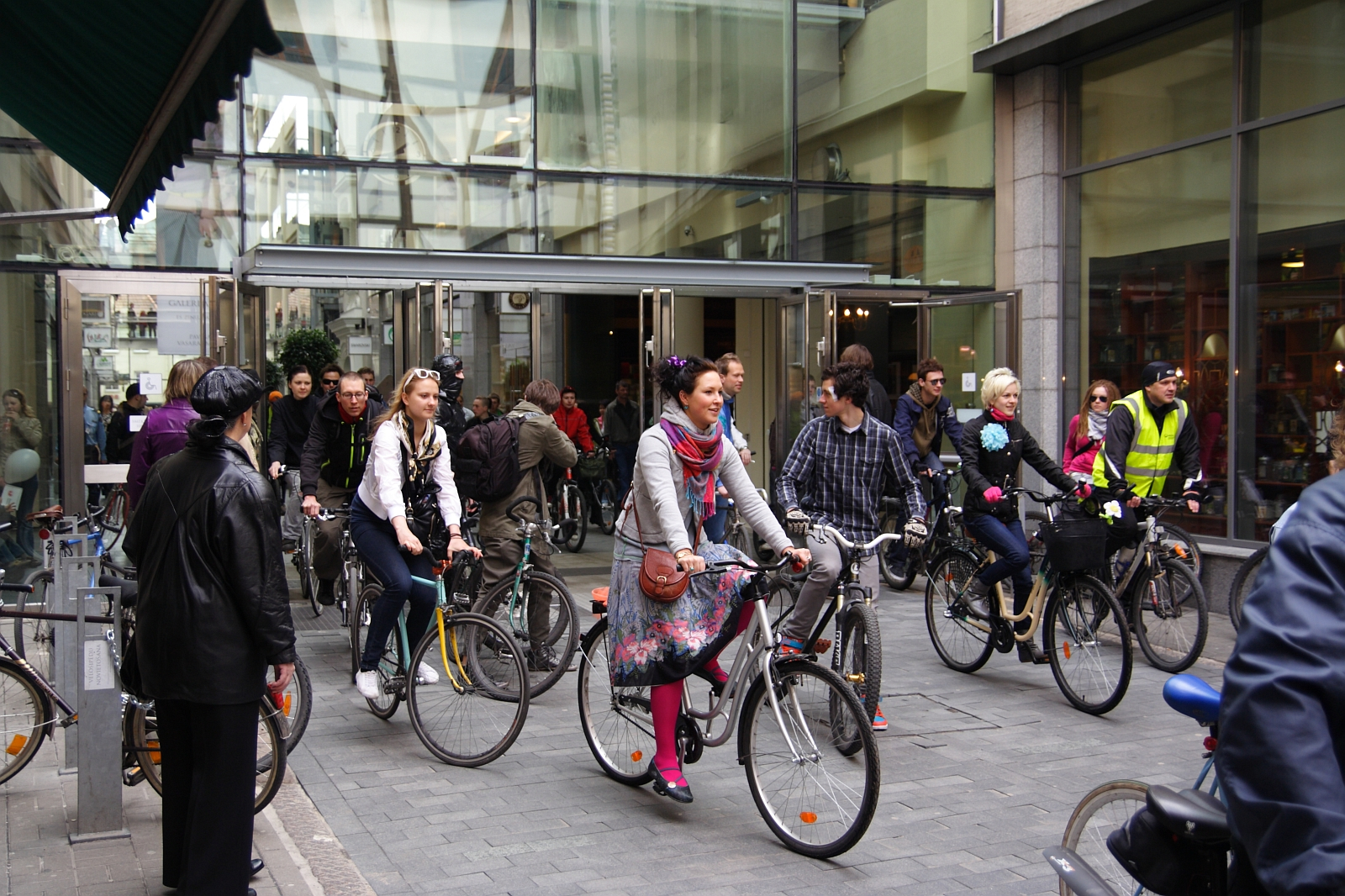 A group of people, dressed in casual and warm clothing, are riding bicycles through a glass-covered pedestrian area during Kritiska Masa 2010. Some cyclists wear helmets, and one person is dressed in a reflective yellow vest. The background shows a modern building with glass windows and doors, and various pedestrians are visible.