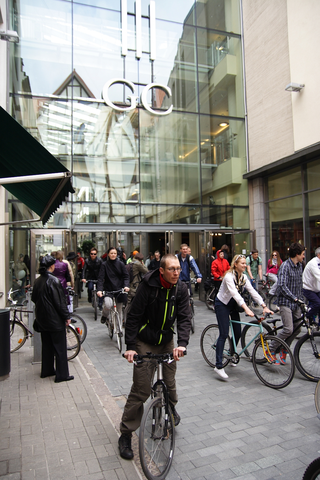 A group of cyclists are riding their bikes on a cobblestone path in front of a modern glass building with the letters "GC" displayed above the entrance. The lively scene resembles the Kritiska masa event from 2010, as pedestrians in various attires watch and add to the bustling atmosphere.