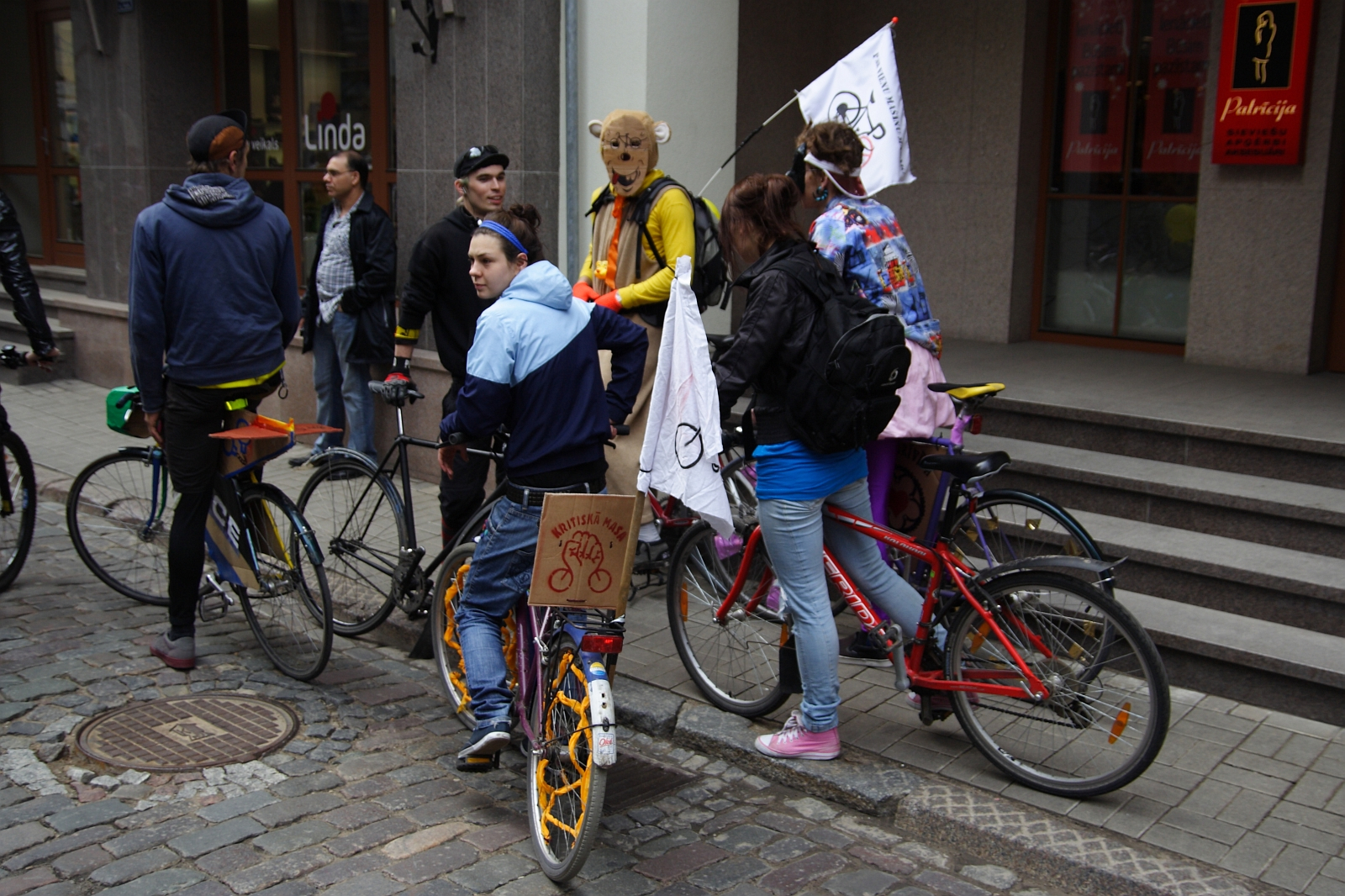 A group of people are gathered on a cobblestone street with their bicycles. One person, wearing a bear costume and holding a flag, stands out. The flags on two of the bicycles display a bicycle symbol. They appear to be participating in the 2010 Kritiska Masa bike event or rally near a building entrance with stairs.