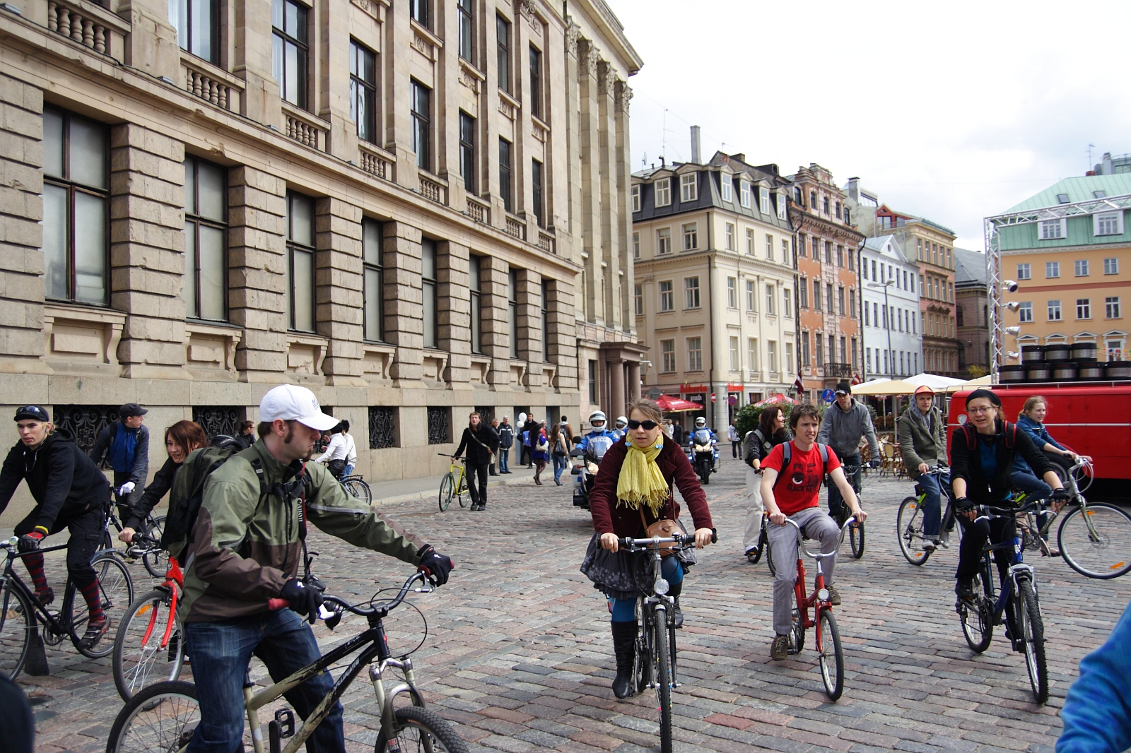 A group of people ride bicycles on a cobblestone street in an urban setting with historic buildings, reminiscent of a 2010 Kritiska masa event. Some cyclists wear helmets and casual clothing, while others don scarves and sunglasses. The sky is partly cloudy, and more pedestrians and cyclists are near a red vendor cart in the background.