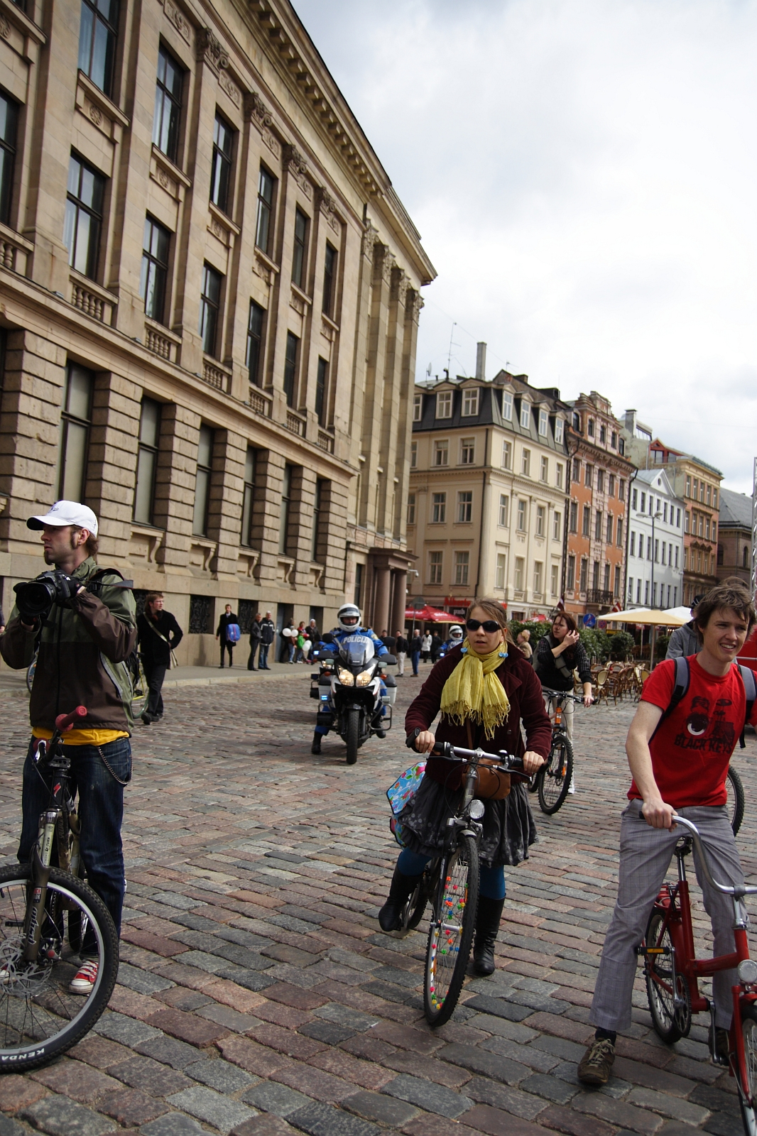 A group of cyclists, part of the 2010 Kritiska Masa, ride on a cobblestone street lined with historic buildings. One cyclist in a yellow scarf and brown jacket rides in the center, while another in a red shirt is on the right. A police officer on a motorcycle and a few pedestrians are in the background. The sky is overcast.
