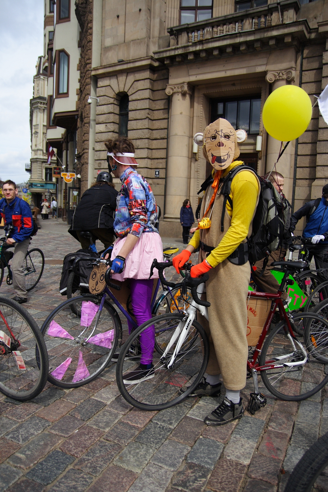 Two cyclists, dressed in colorful and quirky outfits, stand with their bikes in a cobblestone plaza in front of an old building. One cyclist wears a tiger mask, yellow jacket and holds a yellow balloon. The other is clad in a mismatched ensemble with magenta pants. More cyclists from the 2010 Kritiska Masa gather in the background.