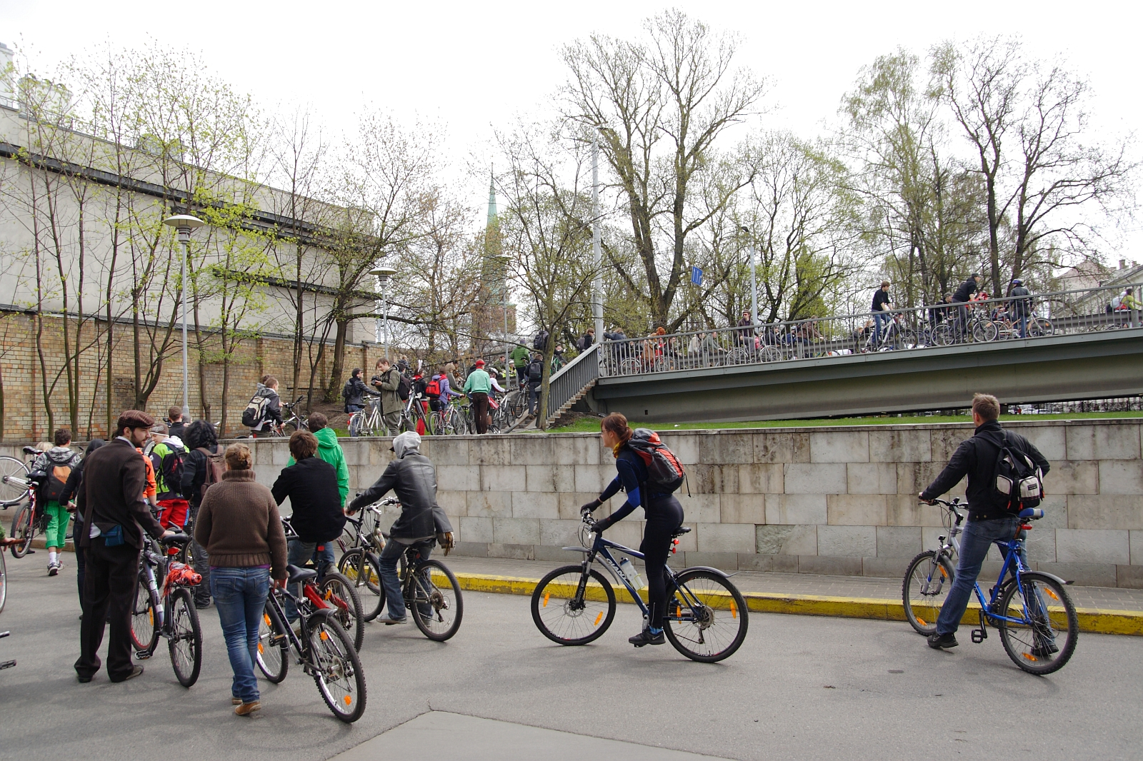 A group of people on bicycles are riding and walking in an urban setting near a gray bridge. The background features leafless trees and a brick building. Some cyclists head up a ramp while others remain at the bottom, evoking memories of the 2010 Kritiska masa cycling event or gathering.