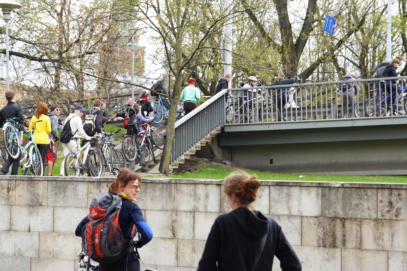 A group of people with bicycles are gathered on and around a pedestrian bridge in an urban park area. Some individuals are walking their bikes up a staircase, while others stand chatting. Trees with early spring foliage surround the scene, embodying the spirit of Kritiska masa. Two women in the foreground converse.