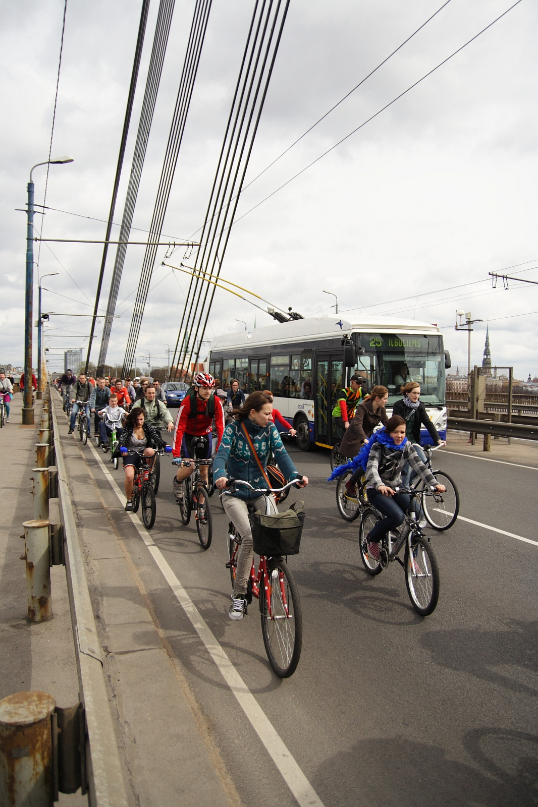 Cyclists ride on a busy bridge next to a moving bus and cars, reminiscent of Kritiska masa 2010. The group includes men, women, and children, some wearing helmets and casual clothing. Overhead wires and streetlights line the bridge, and the sky is overcast. The scene conveys active transportation amidst urban traffic.