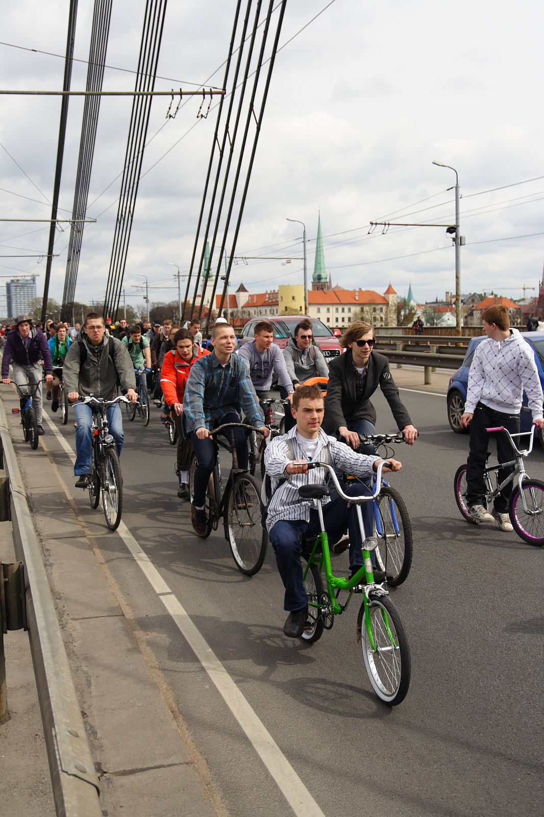 A large group of people, mostly young adults, are riding bicycles along a designated bike lane on a bridge. The cyclists appear to be participating in an organized event akin to the Kritiska masa of 2010. Overhead wires and city buildings with red rooftops and steeples are visible in the background under a cloudy sky.