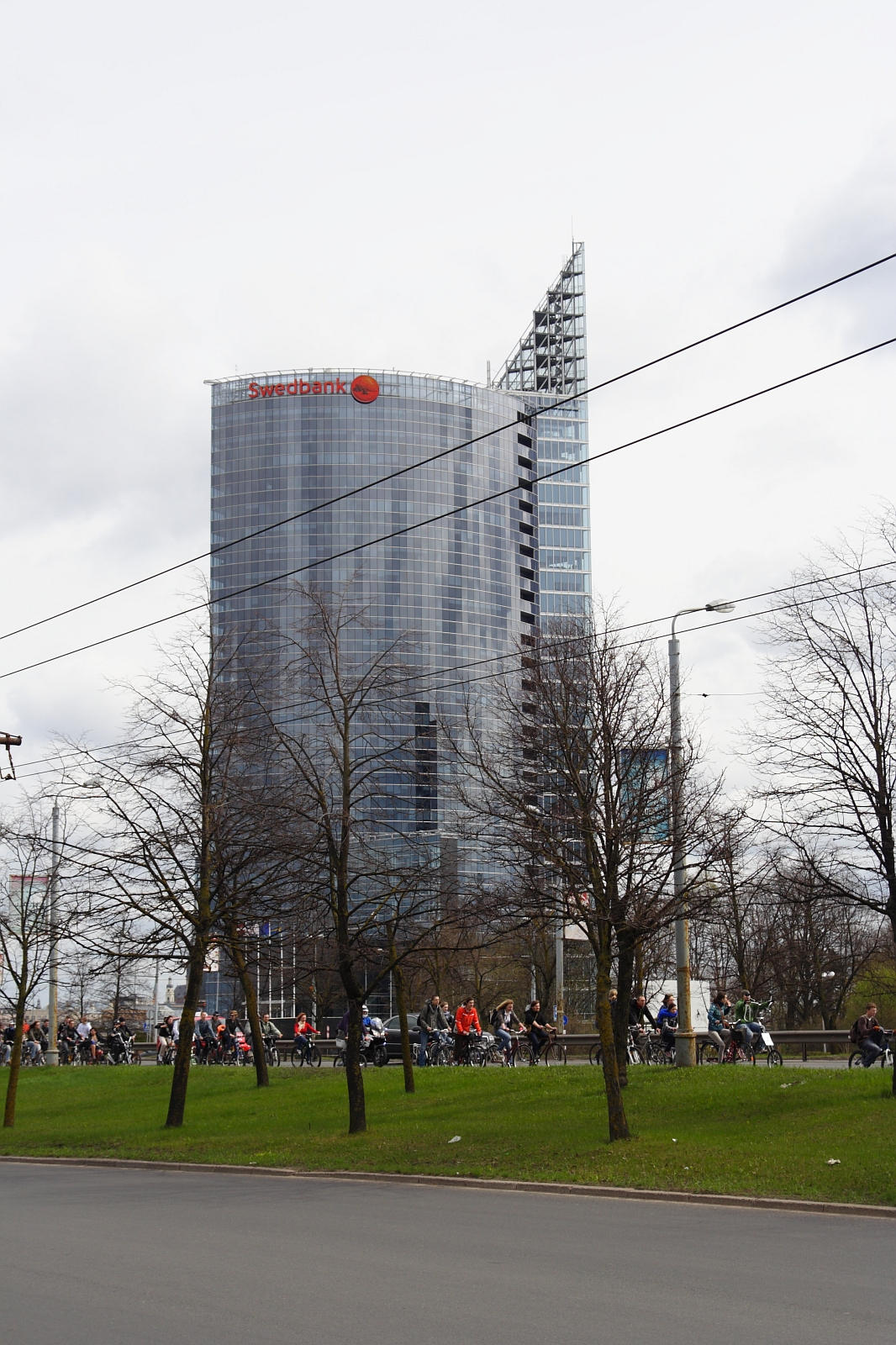 A modern glass skyscraper with the red "Swedbank" logo on top is in the background. In the foreground, a large group of cyclists, part of the Kritiska masa movement, rides along a road lined with leafless trees. Overhead power lines stretch across the cloudy sky in this 2010 urban scene.