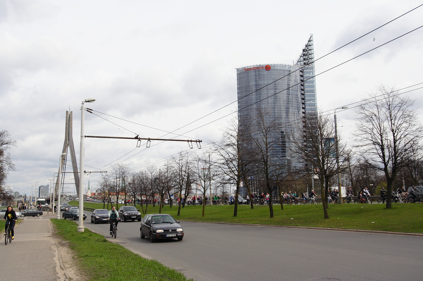 A cityscape featuring a tall glass skyscraper in the background with a distinctive angular design at the top. In the foreground, there is a busy street with cars, cyclists from Kritiska masa, and pedestrians. A modern bridge with cables spans the road, and leafless trees line the street under a cloudy sky, evoking 2010 vibes.