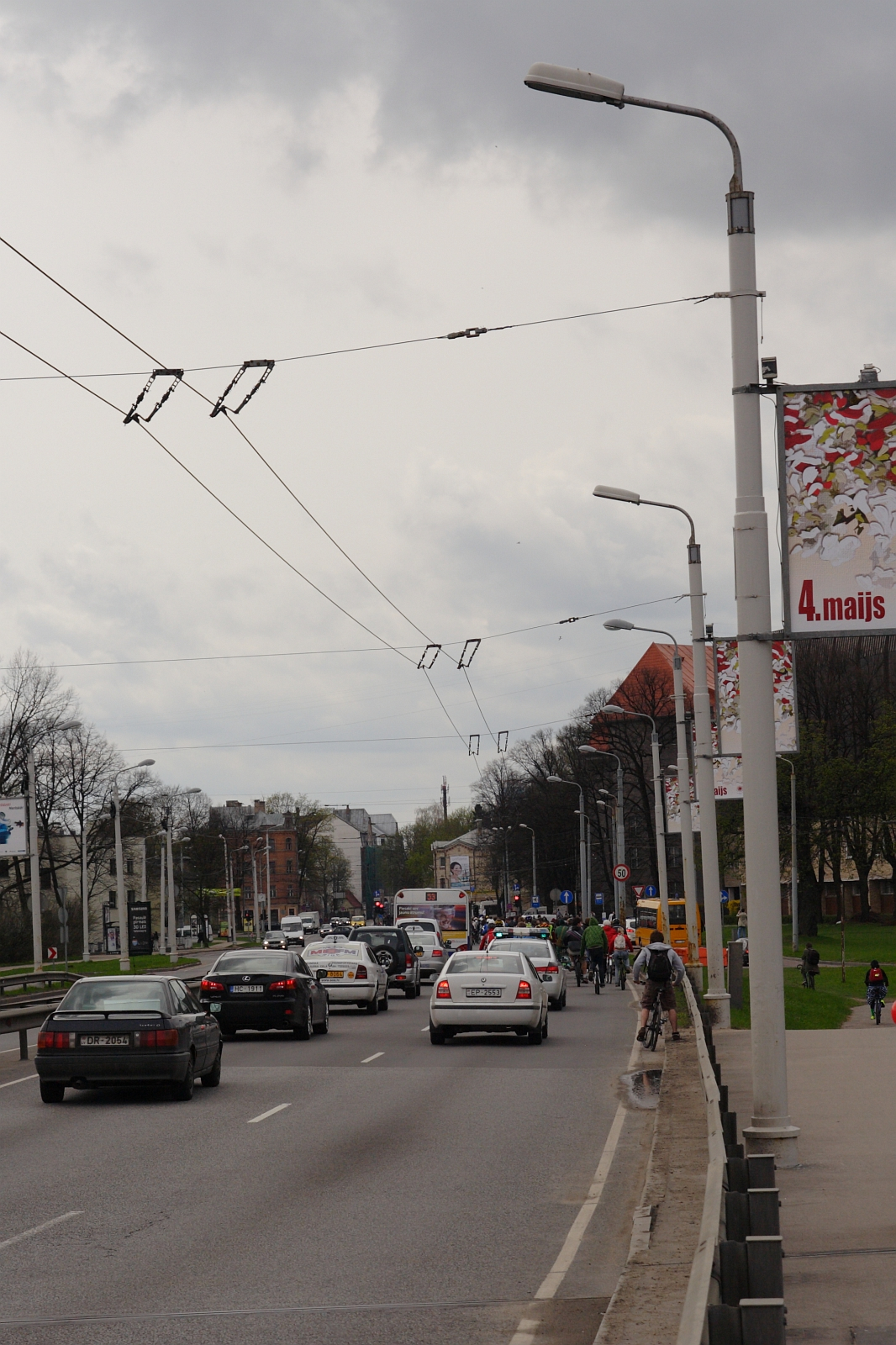 A busy urban street with several cars and a bicyclist, reminiscent of Kritiska Masa 2010, dominates the foreground. Tall lampposts and banners, one reading "4.maijs," line the street. Buildings and trees are visible in the background under a cloudy sky, while overhead power lines crisscross above.