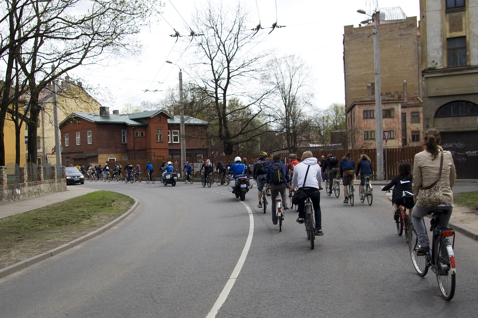 A large group of cyclists ride together on a curved urban street, surrounded by buildings and leafless trees, reminiscent of the 2010 Kritiska masa events. The cyclists range in age and attire, indicating casual participation. A motorcyclist follows behind them. The day appears overcast, and the street has minimal vehicular traffic.