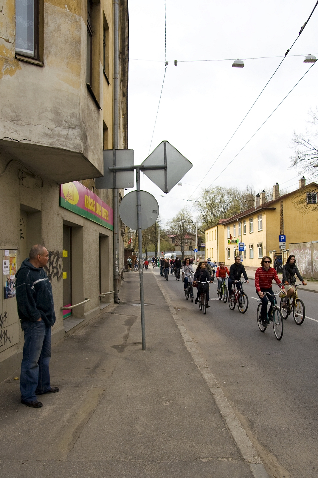 A man stands on the sidewalk near an old building with graffiti and a sign reading "Kinga-Linija" in a foreign language. Multiple cyclists, like those from the Kritiska masa 2010 event, ride down a narrow street under cloudy skies. The street is lined with more old buildings and street signs, and there is a small crowd visible in the distance.
