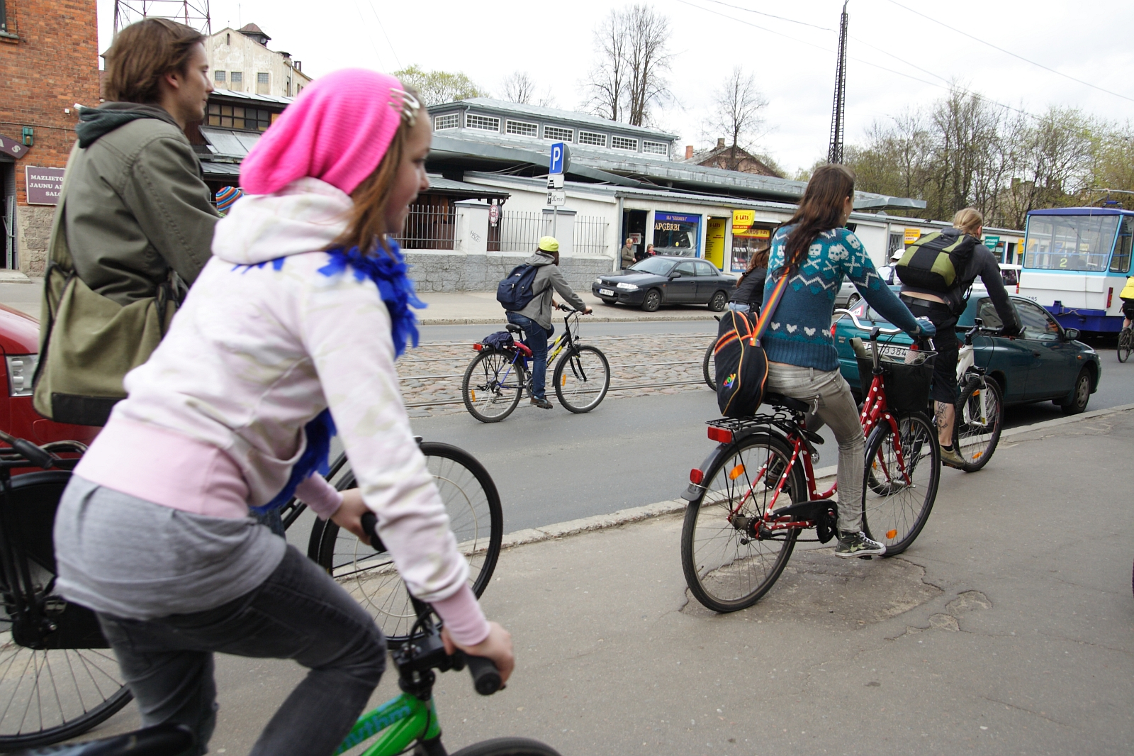 People of various ages are riding bicycles down a city street, reminiscent of a Kritiska Masa event. The closest cyclist, wearing a pink beanie and white hoodie, is on a green bike. Other cyclists are ahead, including one in a teal sweater with a backpack. Buildings, parked cars, and trees line the background.