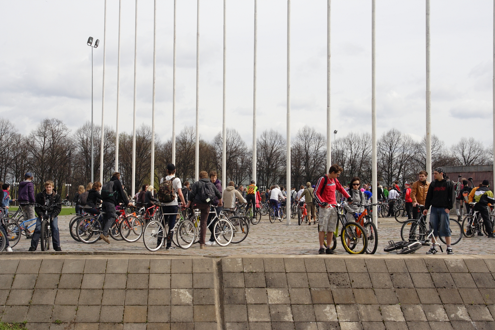 A bustling outdoor scene captures numerous people standing around bicycles, reminiscent of Kritiska Masa 2010. The individuals are in casual clothing, some prepping to ride. The area is paved, and tall flagpoles without flags stand in the background. Bare trees and a cloudy sky frame the setting beautifully.