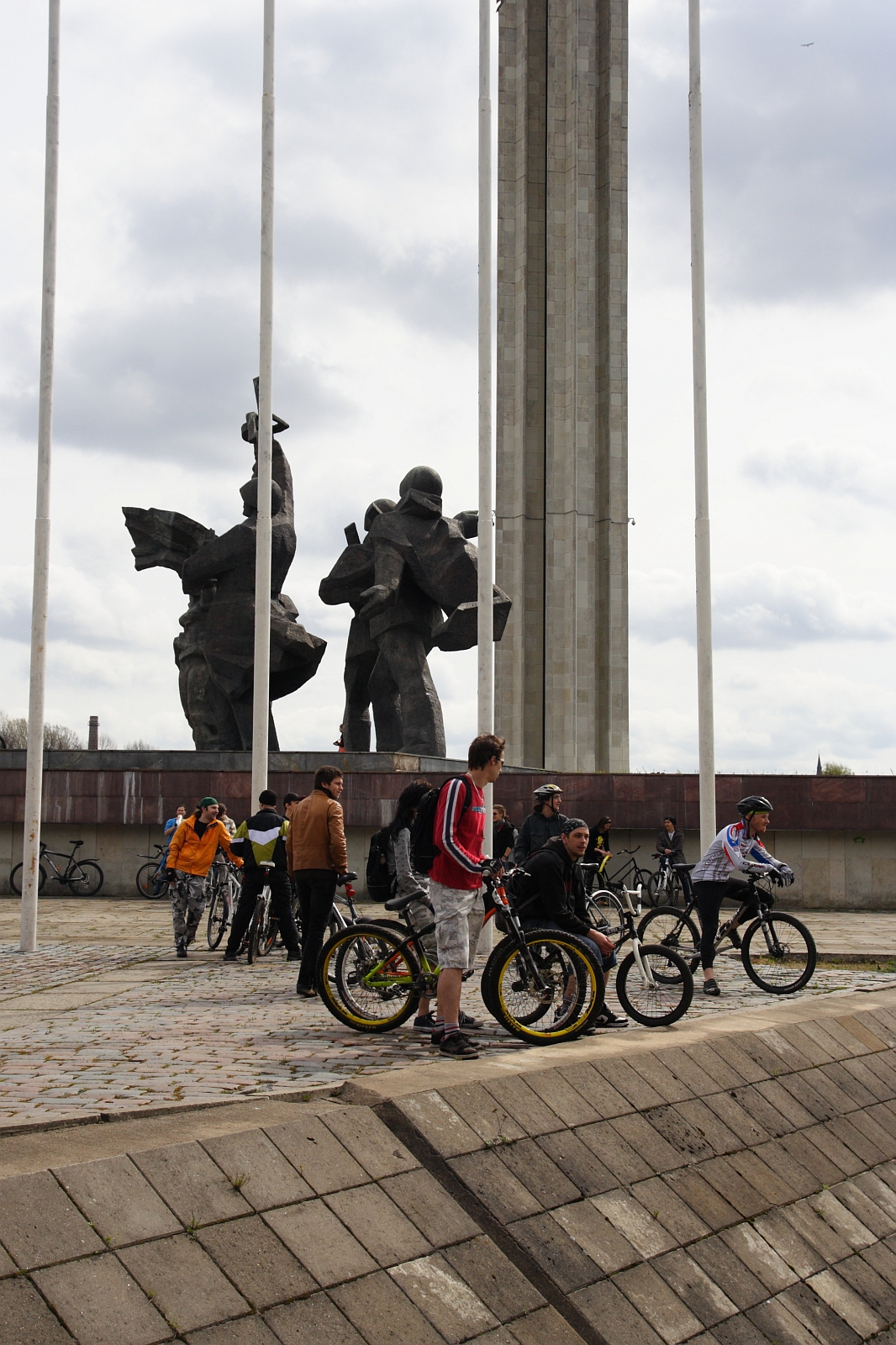 A group of people with bicycles gathers in front of a large, tall monument featuring two abstract statues against a cloudy sky. The scene, reminiscent of Kritiska masa 2010, appears to be in a public plaza with concrete paving and flagpoles. The cyclists are engaged in conversation or preparing their bikes.