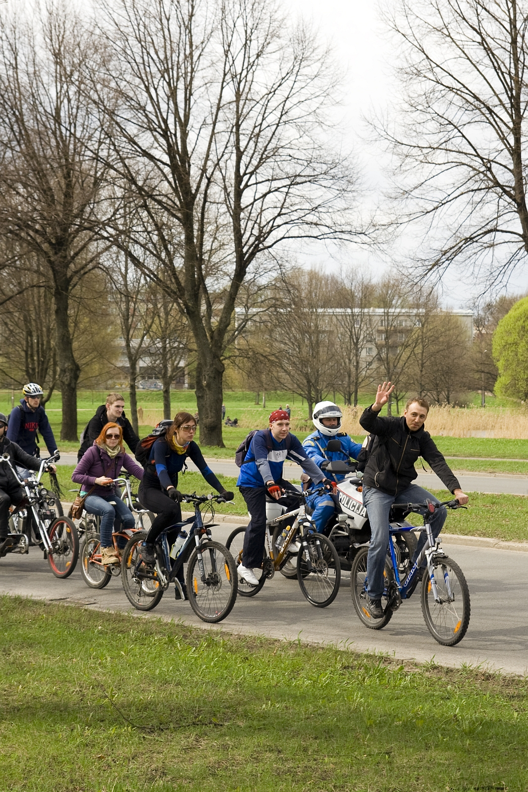 A group of cyclists, including four adults and two children, ride along a paved path in a park during a 2010 Kritiska masa event. The lead male cyclist waves to the camera. Bare trees line the path, and a building with large windows is visible in the background. The sky is overcast, with grass and budding trees surrounding the path.