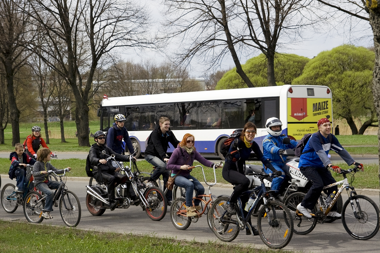 A group of people wearing helmets and casual clothing ride various bicycles and a tandem bike on a paved path beside a grassy area with trees. Behind them, a white bus with purple and yellow accents is in motion. The overcast scene, reminiscent of the 2010 Kritiska masa event, conveys an outdoor activity.