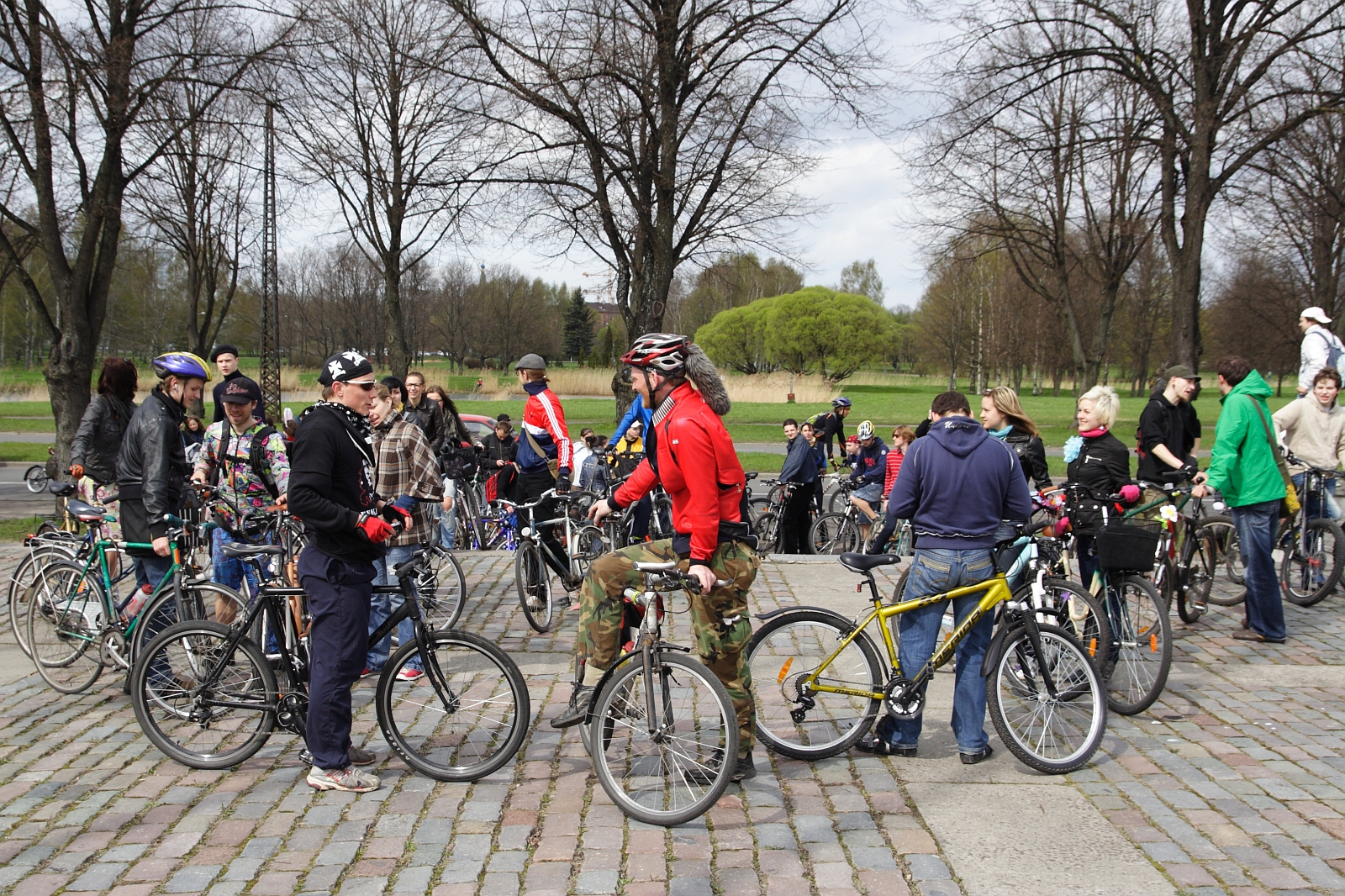 A large group of cyclists gather in a park, chatting and preparing for a ride. They are dressed in casual and sporty clothes, some wearing helmets. The area, paved with cobblestones and surrounded by leafless trees, evokes early spring. The mood is lively and social, resembling a 2010 Kritiska masa event.
