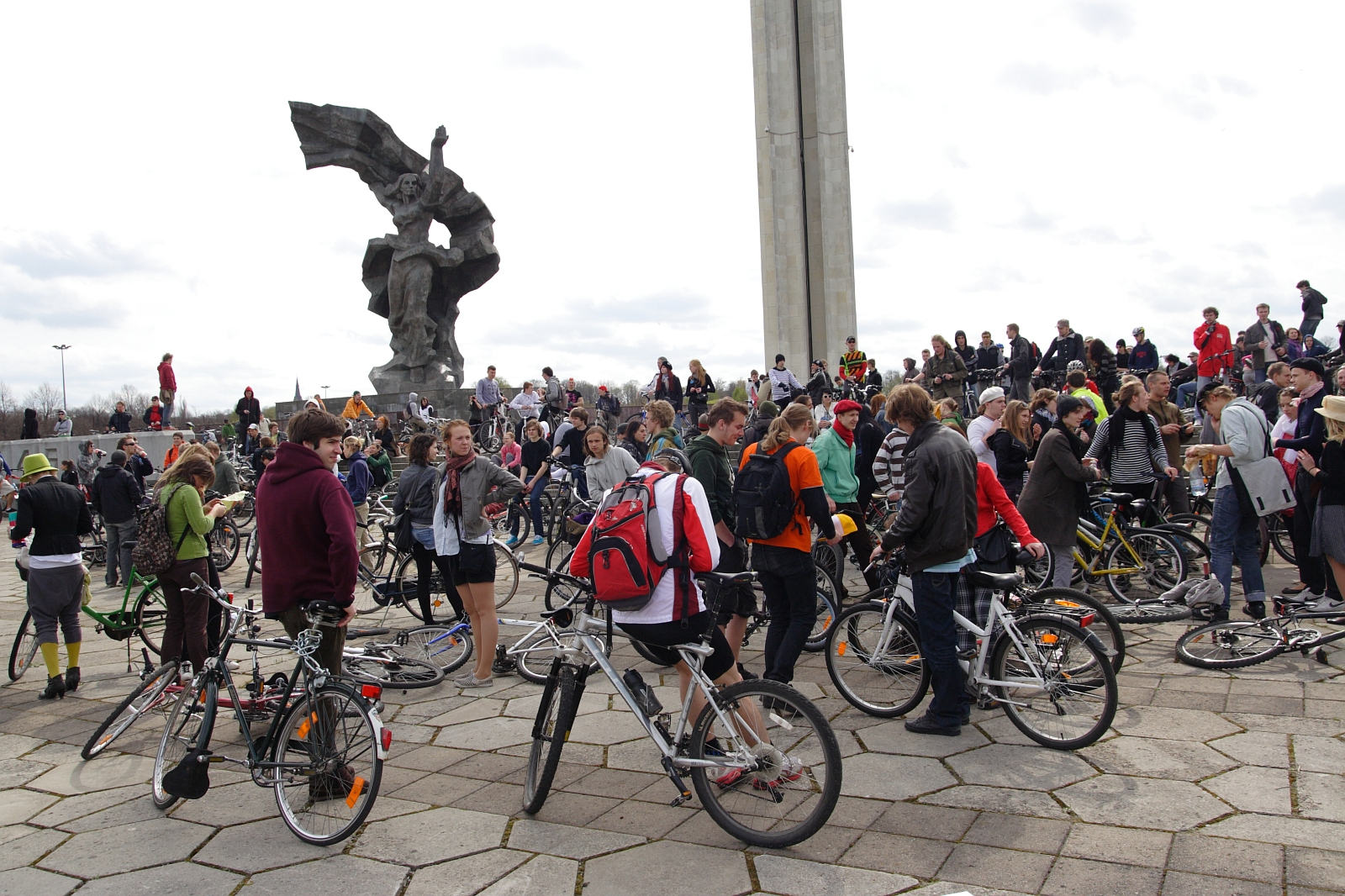 In 2010, a large group of people with bicycles gathered in an open plaza featuring a tall sculpture in the background, reminiscent of a Kritiska masa event. Many individuals are standing and conversing, with some sitting on their bikes. The atmosphere appears lively and social, with a mix of cloudy skies overhead.