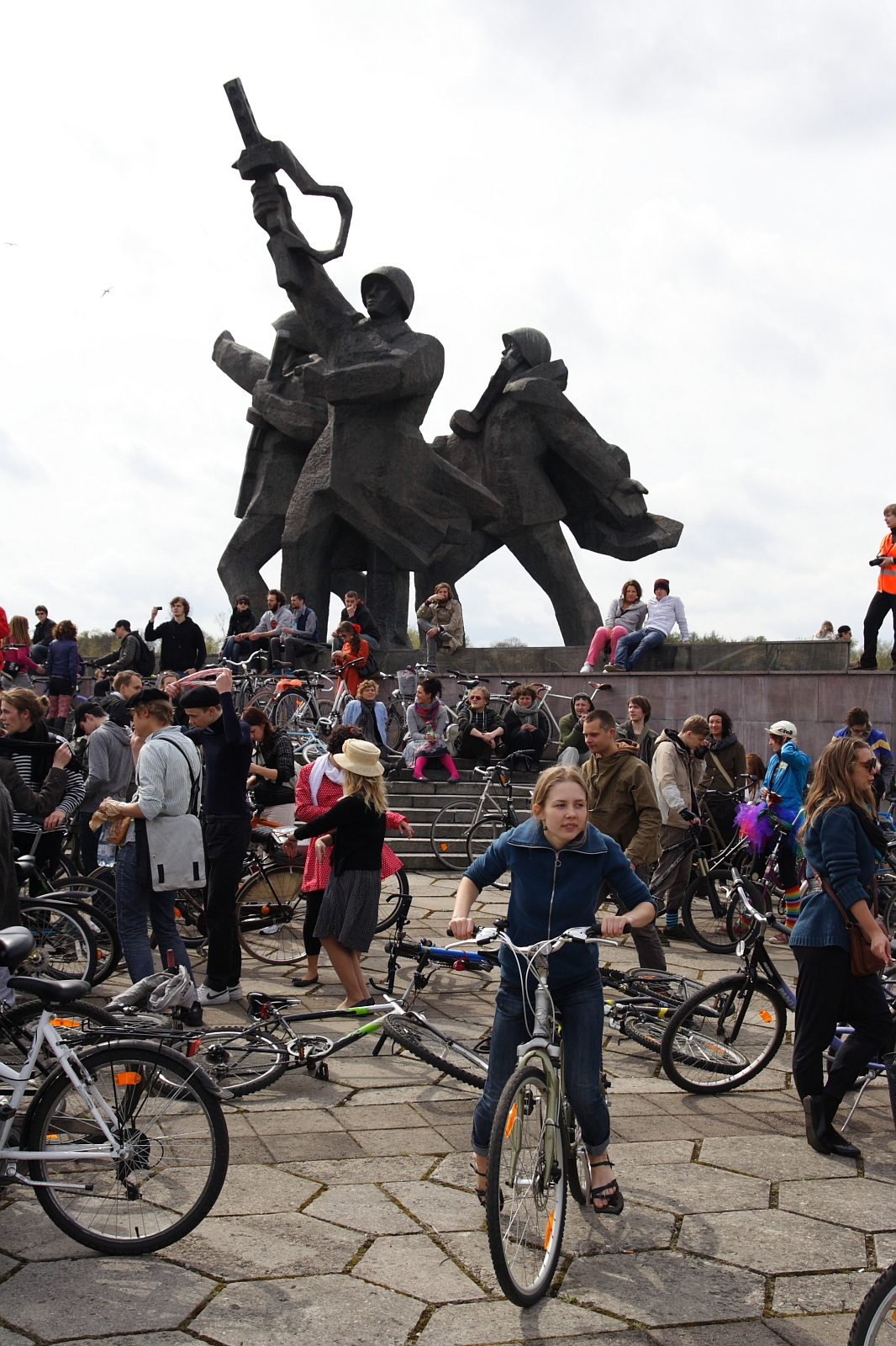 A young girl in a blue jacket rides her bike in front of a crowd gathered around a large, dark, abstract monument depicting several figures holding tools and weapons. On this cloudy day, during Kritiska masa 2010, people of various ages, some with bicycles, gather, sit, and stand around the statue.