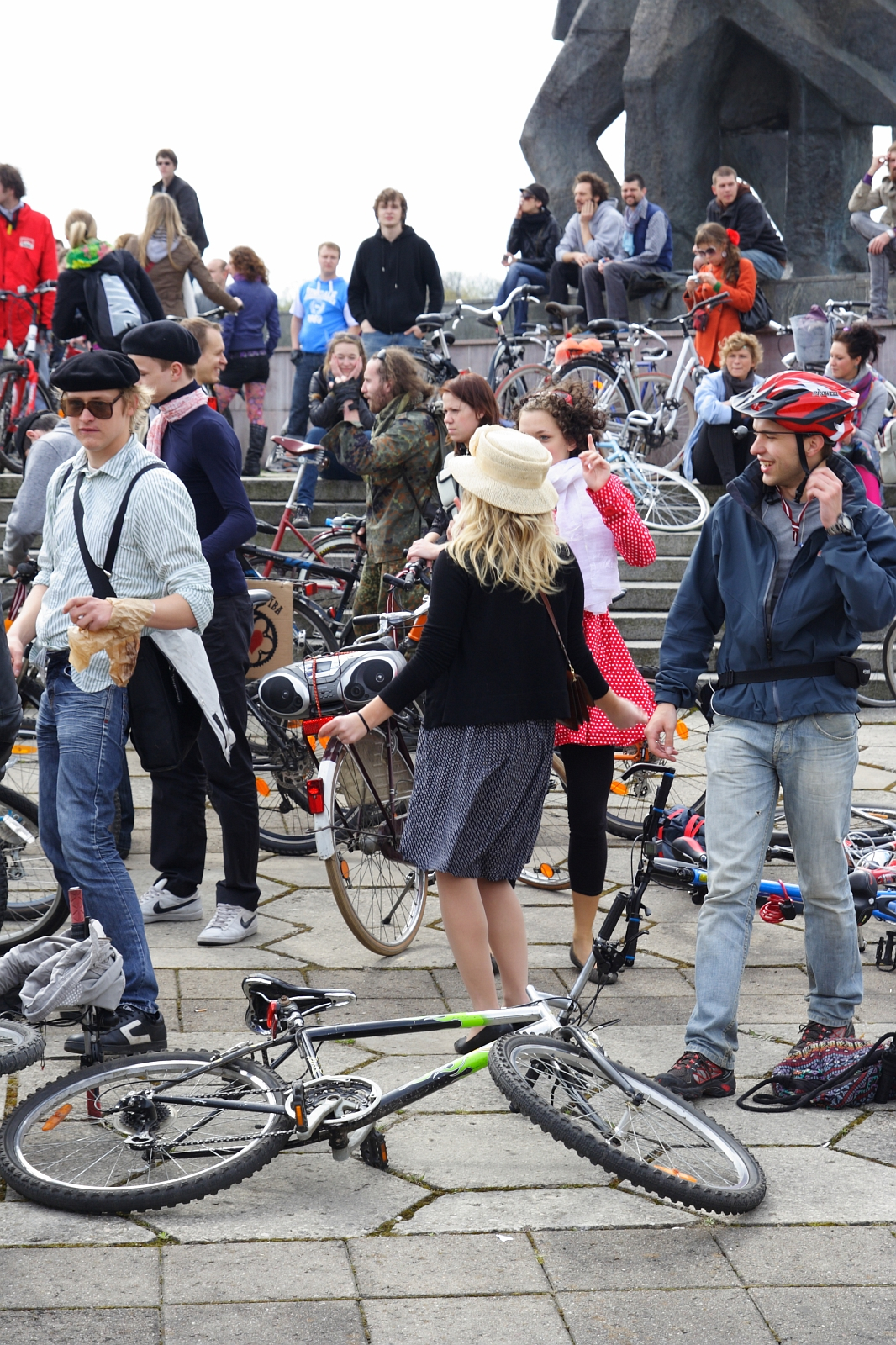 A diverse crowd of people, many with bicycles, gather outdoors on a paved area during the 2010 Kritiska masa event. Some stand while others sit on steps in the background. A woman in a black top and striped skirt holds a bike with a basket. Many wear helmets as they engage in lively conversation, creating a vibrant atmosphere.