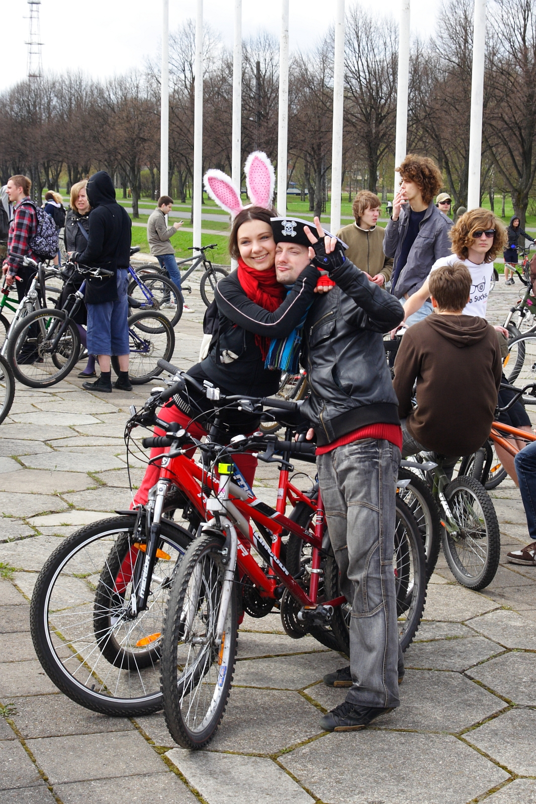 A joyful couple, the woman wearing white bunny ears, pose with their red bicycles at the 2010 Kritiska masa event. The man, dressed in a black leather jacket, flashes a peace sign. Joined by other cyclists and some walkers, they gather outdoors amidst leafless trees. The atmosphere is festive and lively.