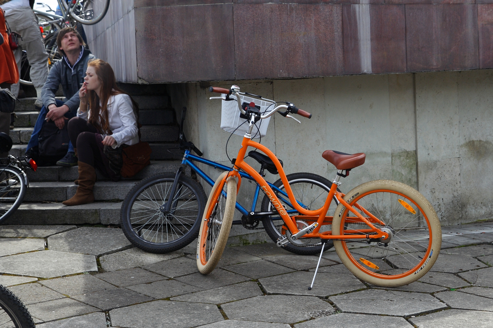 Two people sit on steps near a building, reminiscent of Kritiska masa gatherings. An orange bike with light tan tires and a blue bike rest against the steps. The pavement is irregularly shaped concrete tiles. The seated individuals are engaged in conversation, and one person appears to be looking at something above.