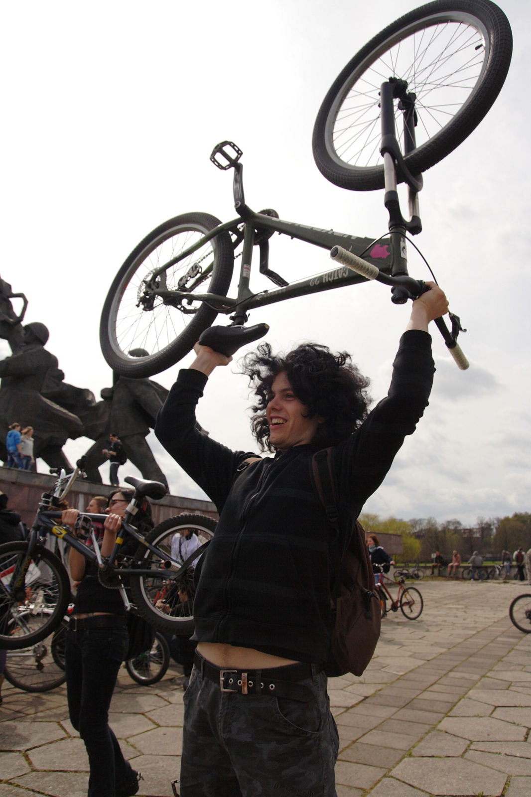 A person with curly hair and a black hoodie lifts a bicycle above their head outdoors during Kritiska Masa 2010. They are smiling, and other people with bicycles are visible in the background near a large stone sculpture. The sky is overcast, and the ground is paved with stone tiles.
