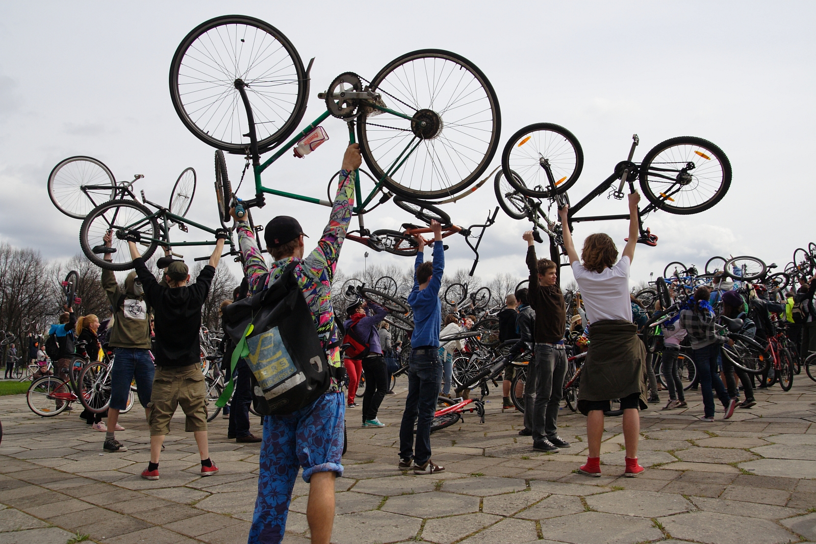 A diverse group of cyclists, part of the 2010 Kritiska masa event, energetically hold their bicycles above their heads in a celebratory manner. They stand on a paved area with a cloudy sky above, demonstrating enthusiasm and community spirit. Trees can be seen in the background.