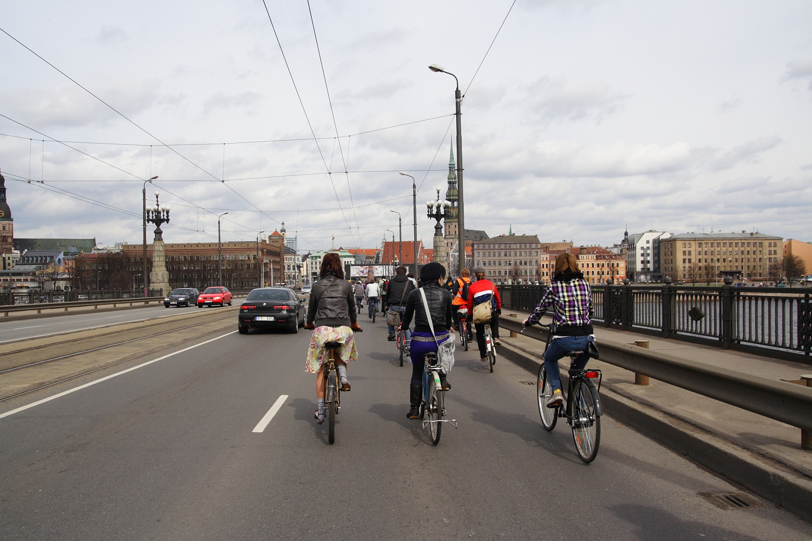 A group of cyclists rides across a wide bridge alongside a single lane of car traffic, under a cloudy sky. The cyclists, part of a Kritiska masa event, are dressed in casual attire. The bridge features tram tracks and lamp posts, with historic buildings and a church steeple visible in the skyline.